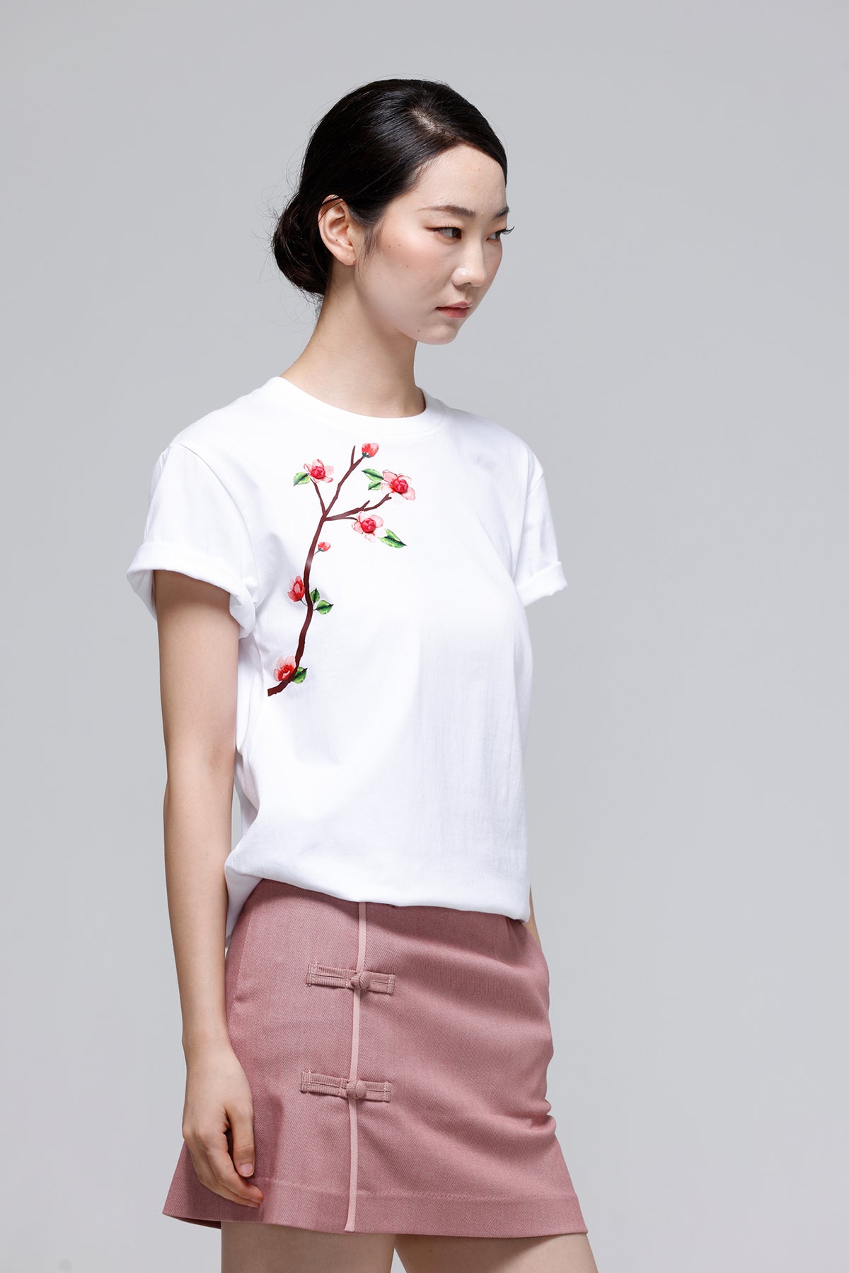 A woman wearing a white short-sleeve crewneck t-shirt with a red and green floral branch embroidery on the left chest, paired with a dusty pink high-waisted mini skirt featuring traditional Chinese frog closures, set against a solid light gray background.