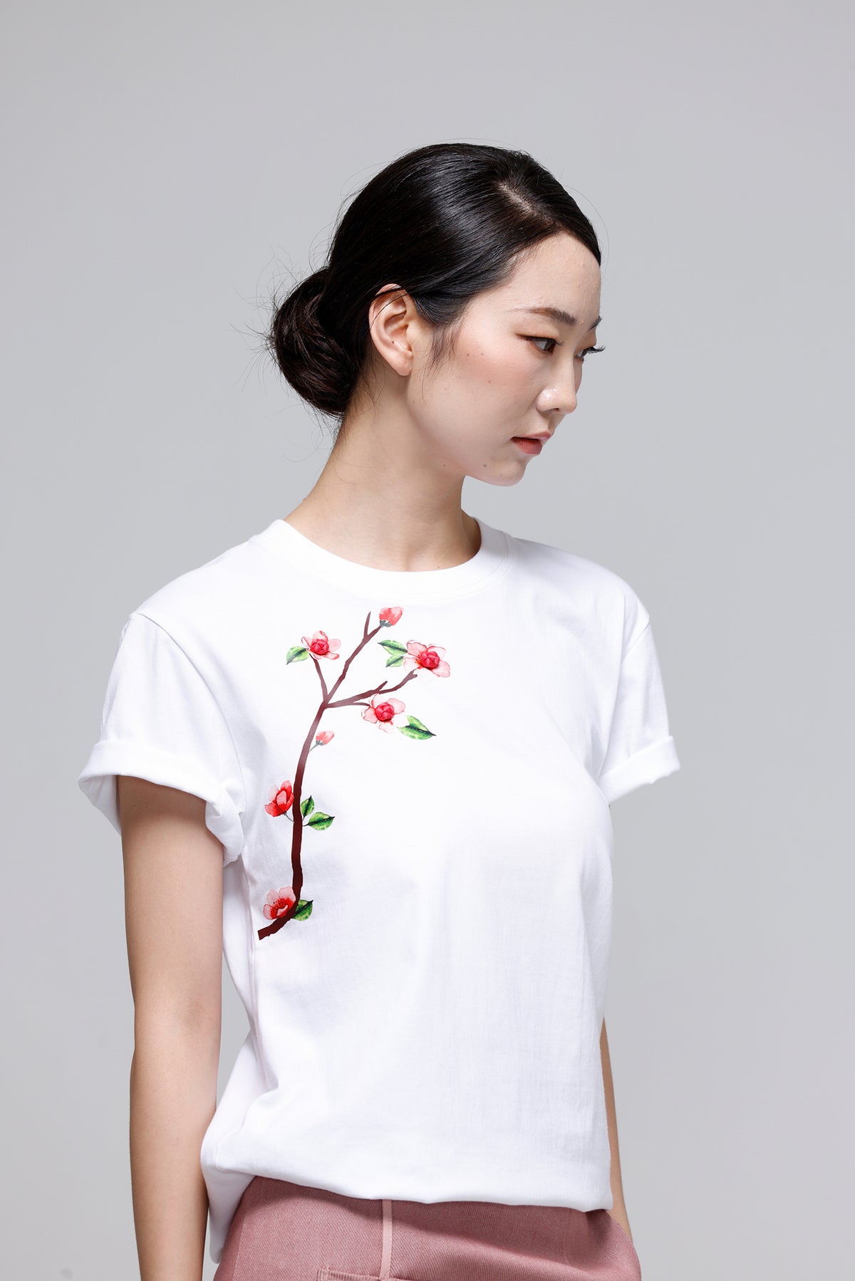 A woman wearing a white short-sleeve crewneck t-shirt with an embroidered cherry blossom branch on the front, paired with dusty rose pants, set against a plain light gray studio background.