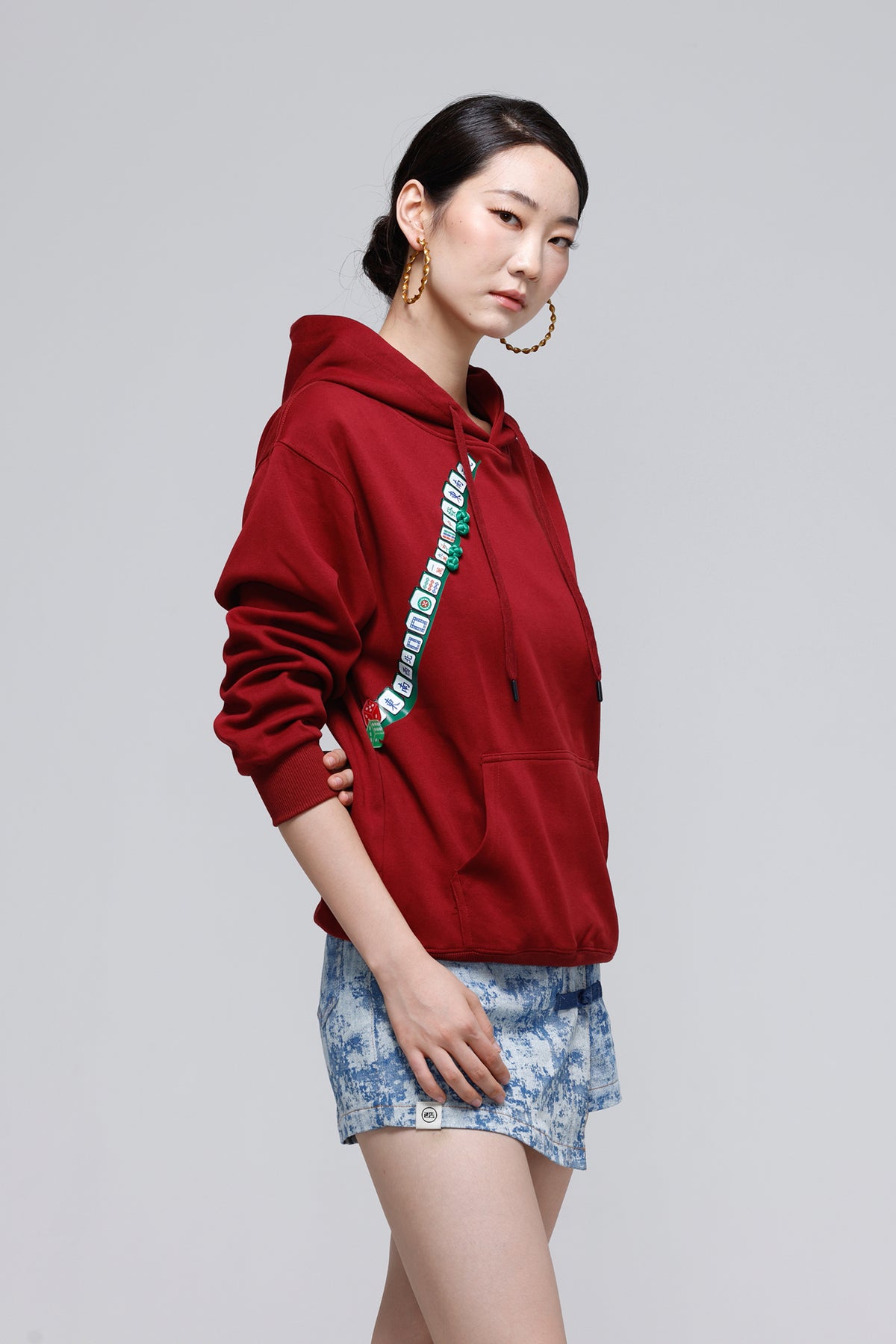 A woman modeling a deep red pullover hoodie with a green and white patterned vertical stripe detail on the front, paired with blue denim shorts, against a solid light gray background.