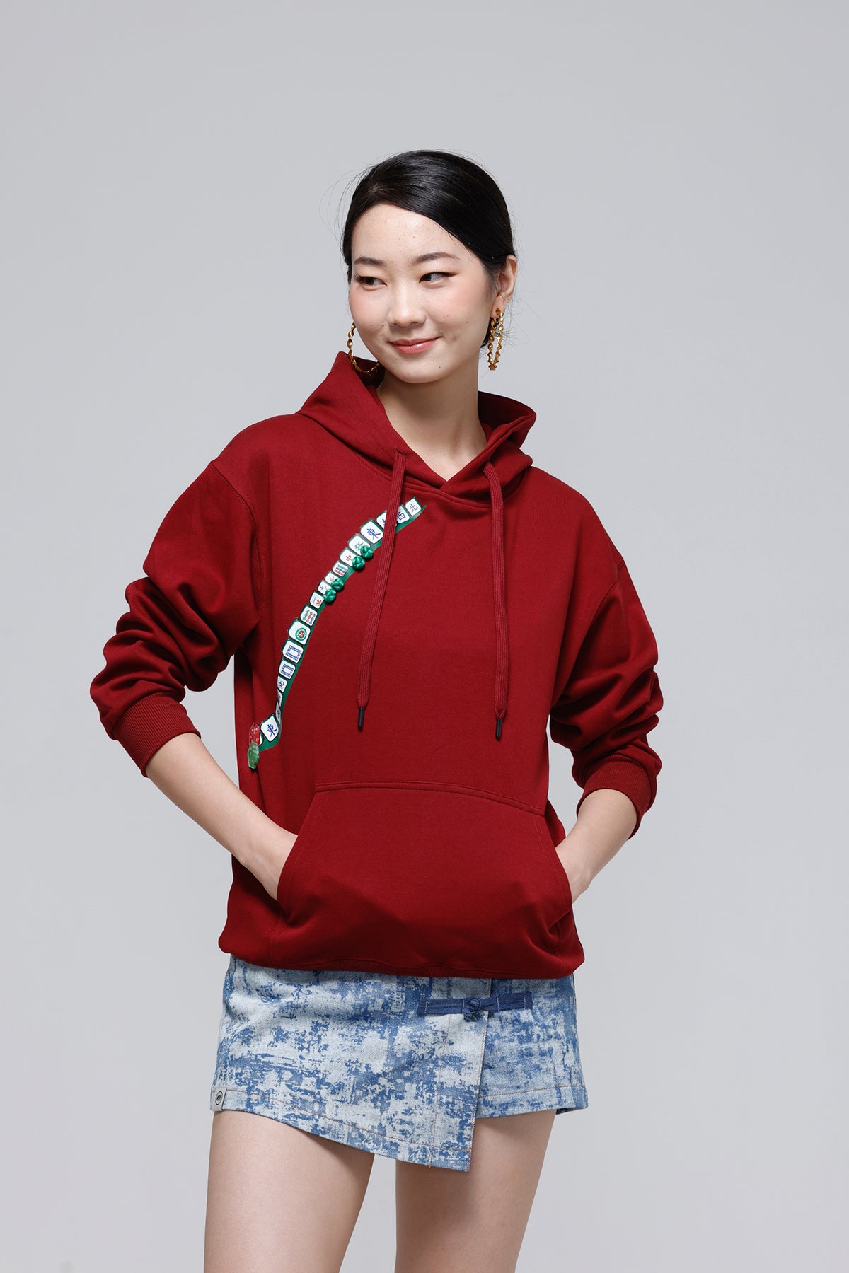 A woman modeling a deep red pullover hoodie with a green and white graphic print along the left chest, paired with blue patterned shorts, against a solid light gray background.