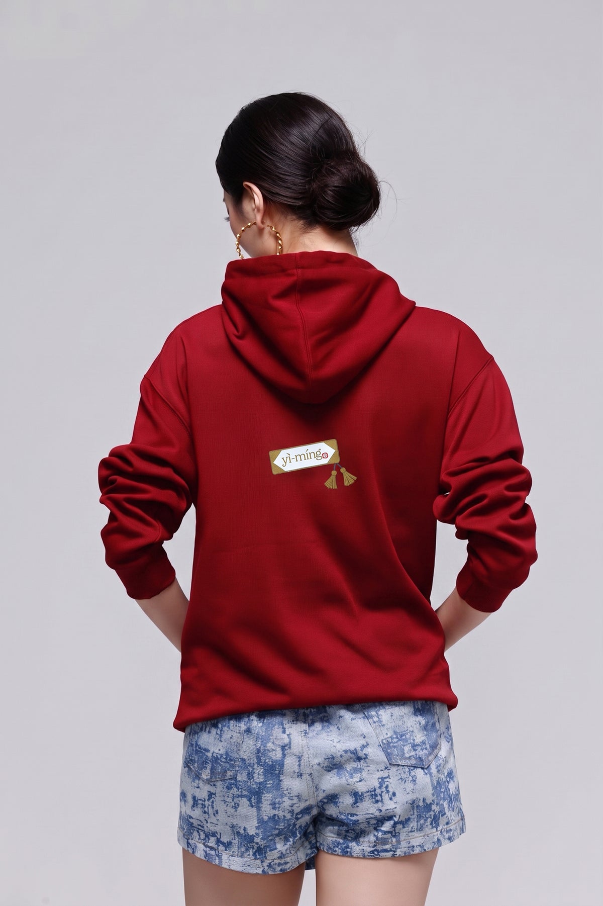 A woman viewed from behind wearing a deep red hoodie with a small gold and white graphic on the back, paired with blue and white patterned shorts, against a solid light gray background.