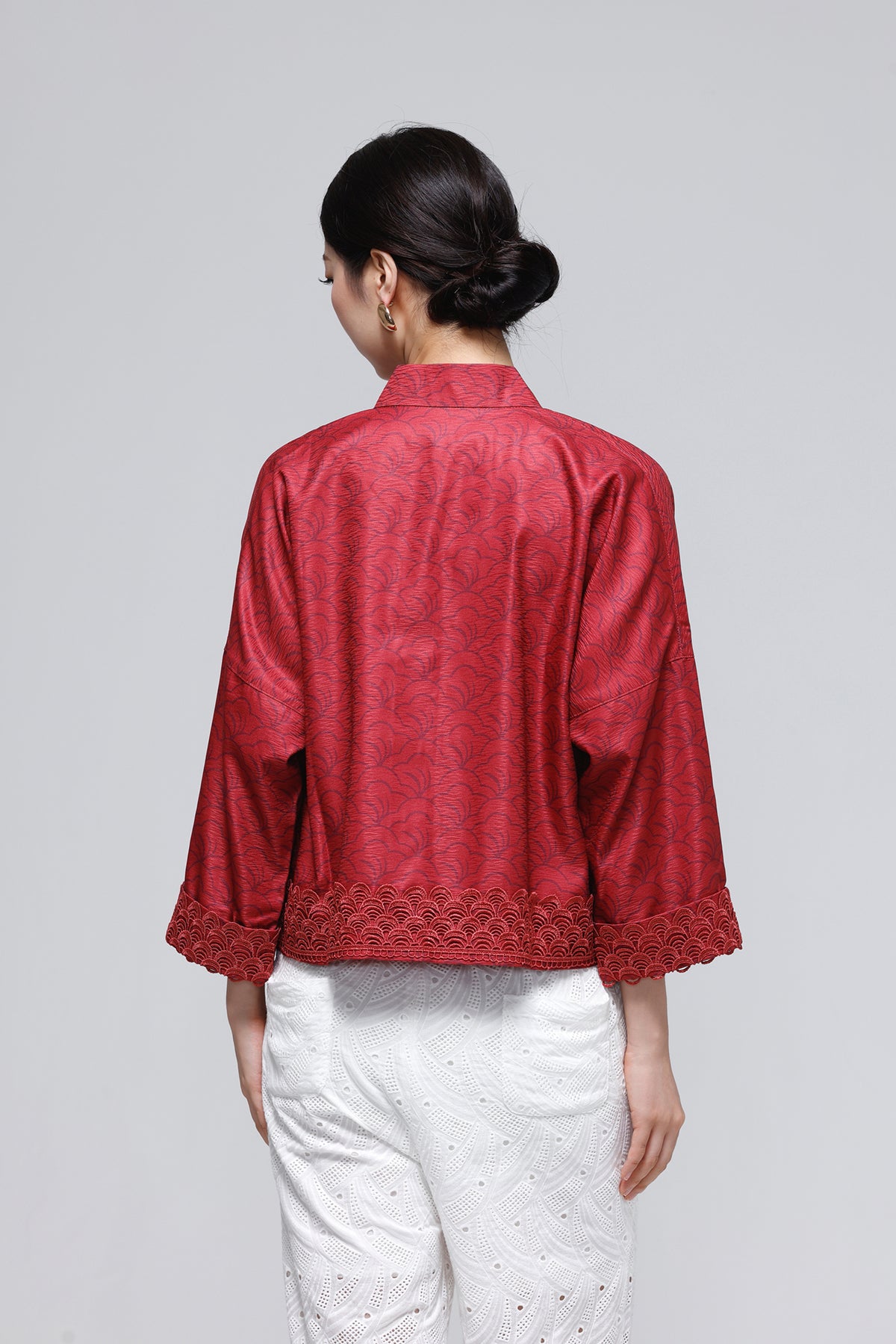 A woman shown from the back wearing a red textured jacket with three-quarter sleeves and white pants, photographed against a solid light gray background.