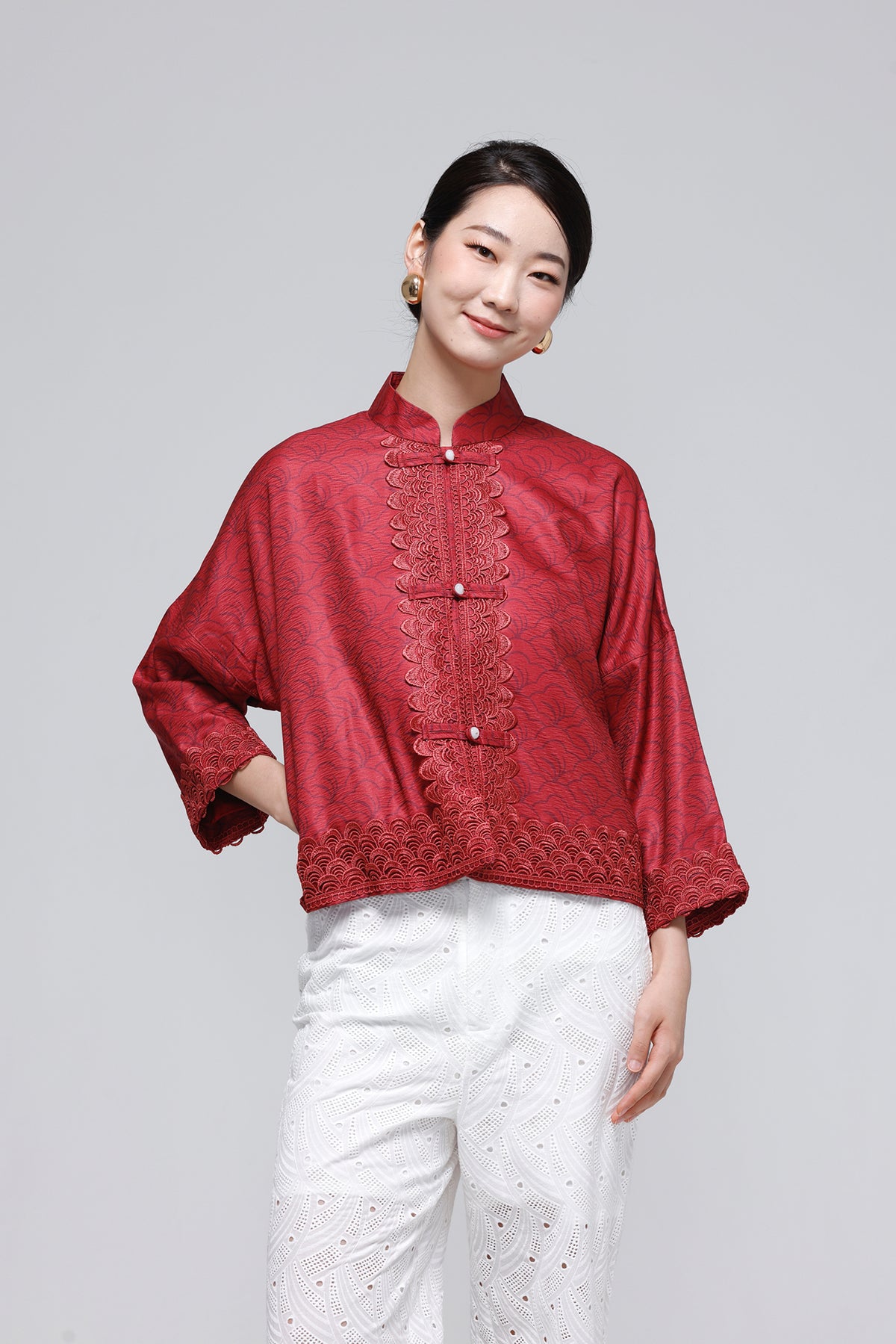 A woman modeling a red textured Chinese-style jacket with lace trim and white pants against a solid gray studio background.