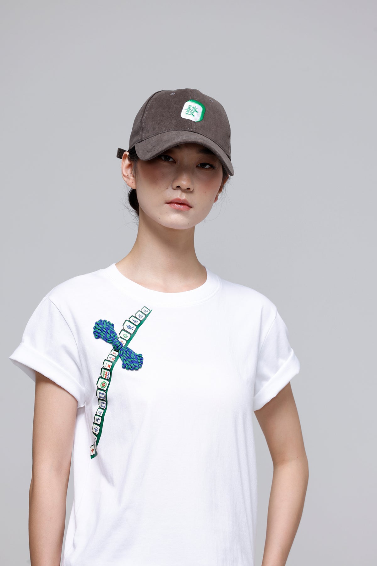 A model wearing a white short-sleeve t-shirt with a blue and green graphic print on the front, paired with a dark brown baseball cap, against a solid gray studio background.