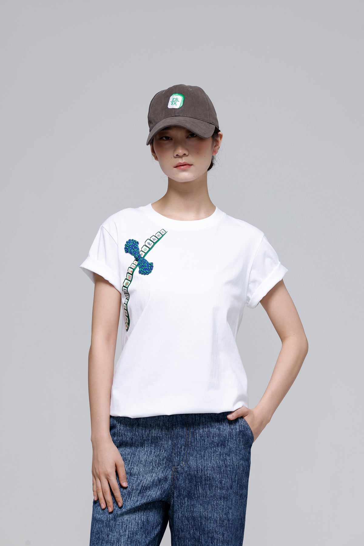 A woman modeling a white short-sleeve crewneck t-shirt with a blue and green graphic print on the left chest, paired with a brown baseball cap and blue jeans, against a solid gray studio background.