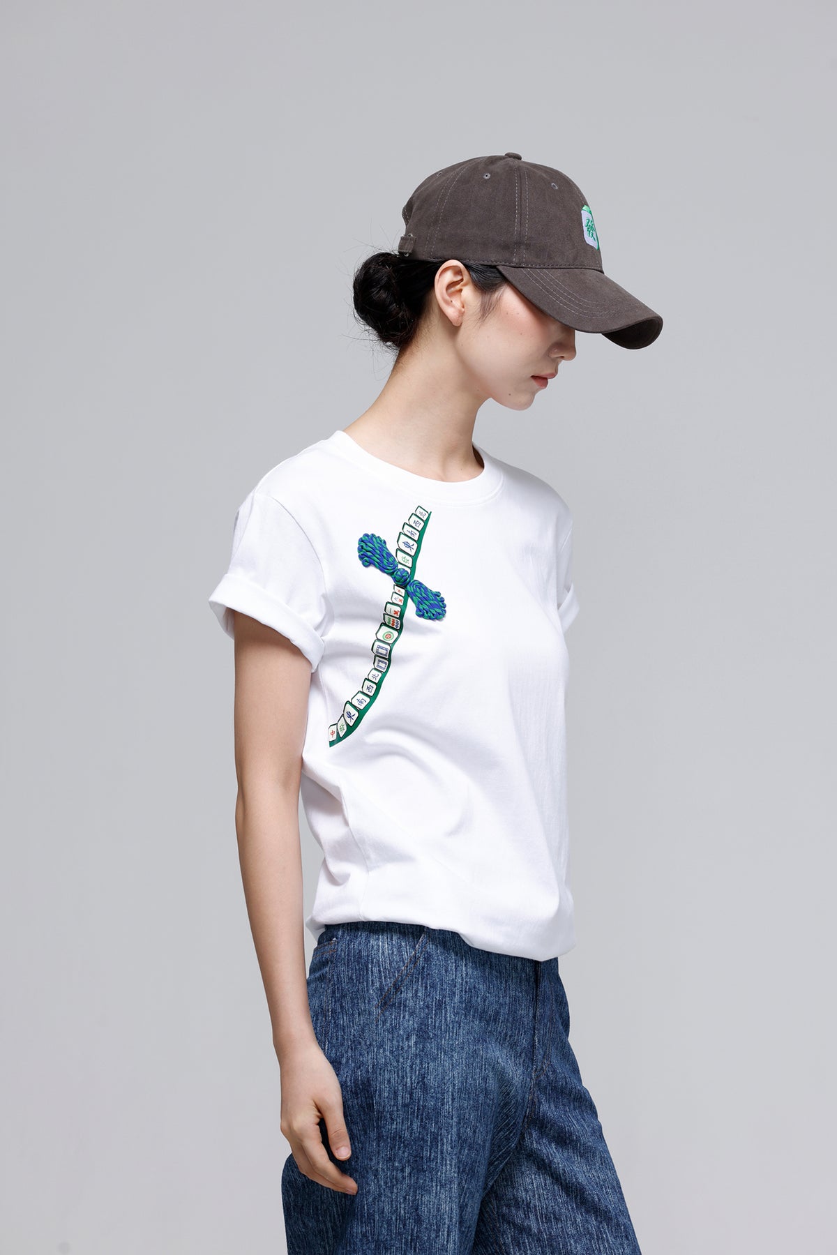A woman wearing a white short-sleeve t-shirt with a blue and green graphic print on the chest, paired with blue jeans and a dark brown baseball cap, photographed against a solid light gray background.