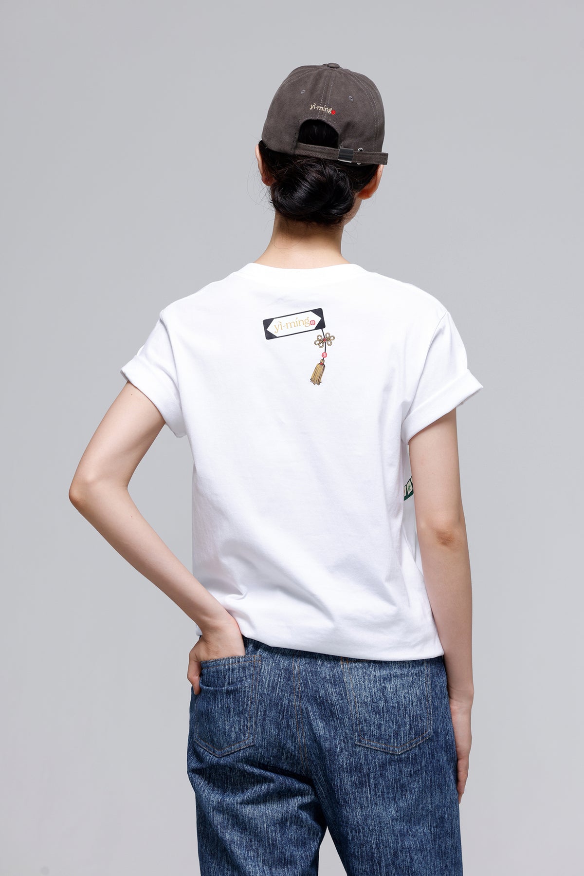 A person shown from the back wearing a white short-sleeve T-shirt with a small graphic print on the upper back, blue jeans, and a dark baseball cap, against a light gray studio background.
