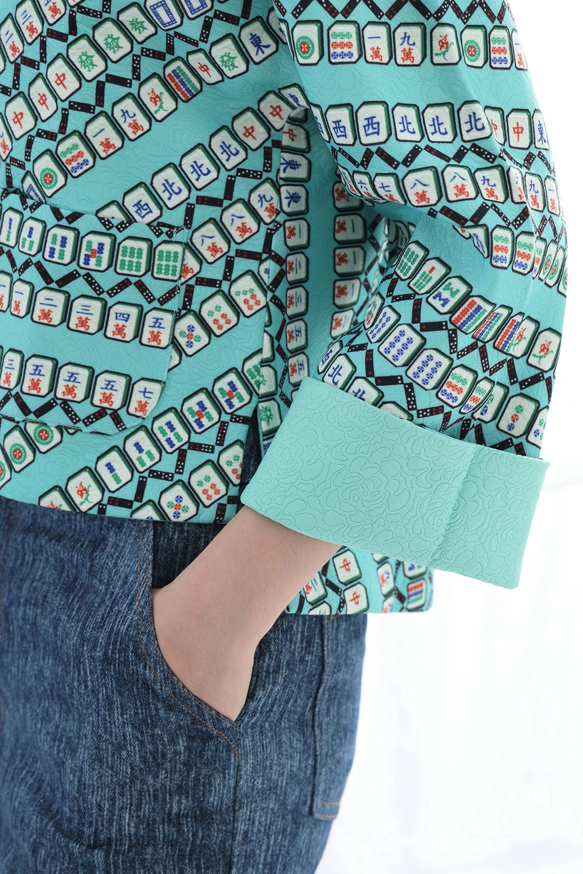 A close-up of a person wearing a mint green long-sleeve blouse with a black and multicolor mahjong tile pattern, paired with blue denim jeans.