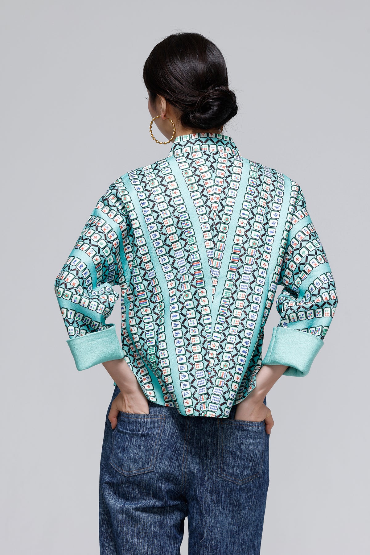 A woman viewed from the back wearing a mint green and black patterned long-sleeve blouse with contrasting solid mint green trim, paired with blue jeans, against a solid gray studio background.