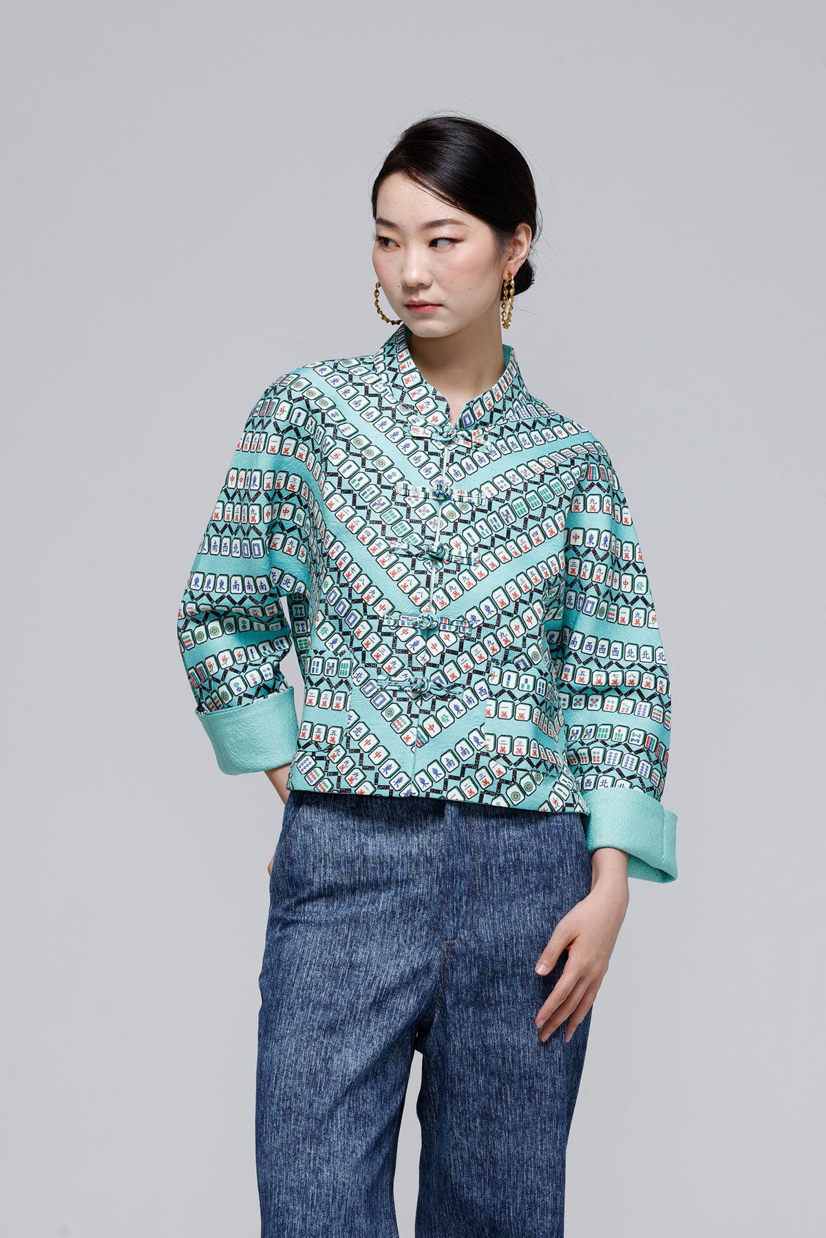 A woman modeling a long-sleeve button-up shirt with a geometric pattern in teal, black, and white, paired with blue jeans, against a solid gray studio background.