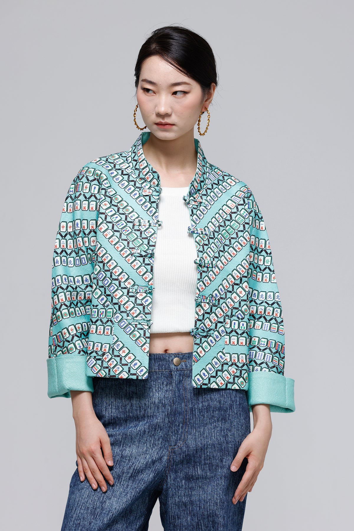 A model wearing a cropped, open-front jacket with a teal and white geometric pattern, styled with a white ribbed top and blue jeans against a solid gray background.