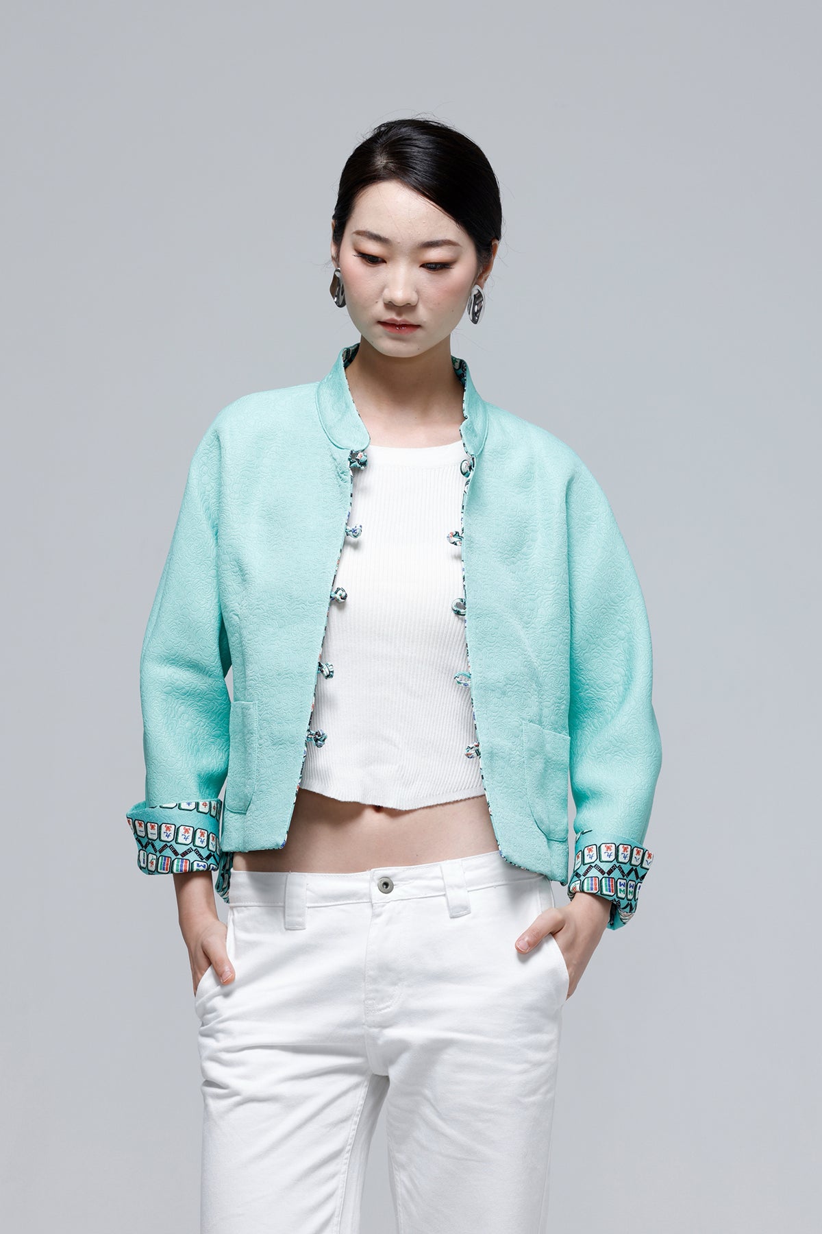 A woman modeling a light turquoise cropped jacket with decorative silver-tone buttons and patterned cuffs, worn over a white crop top and white pants, against a plain light gray studio background.