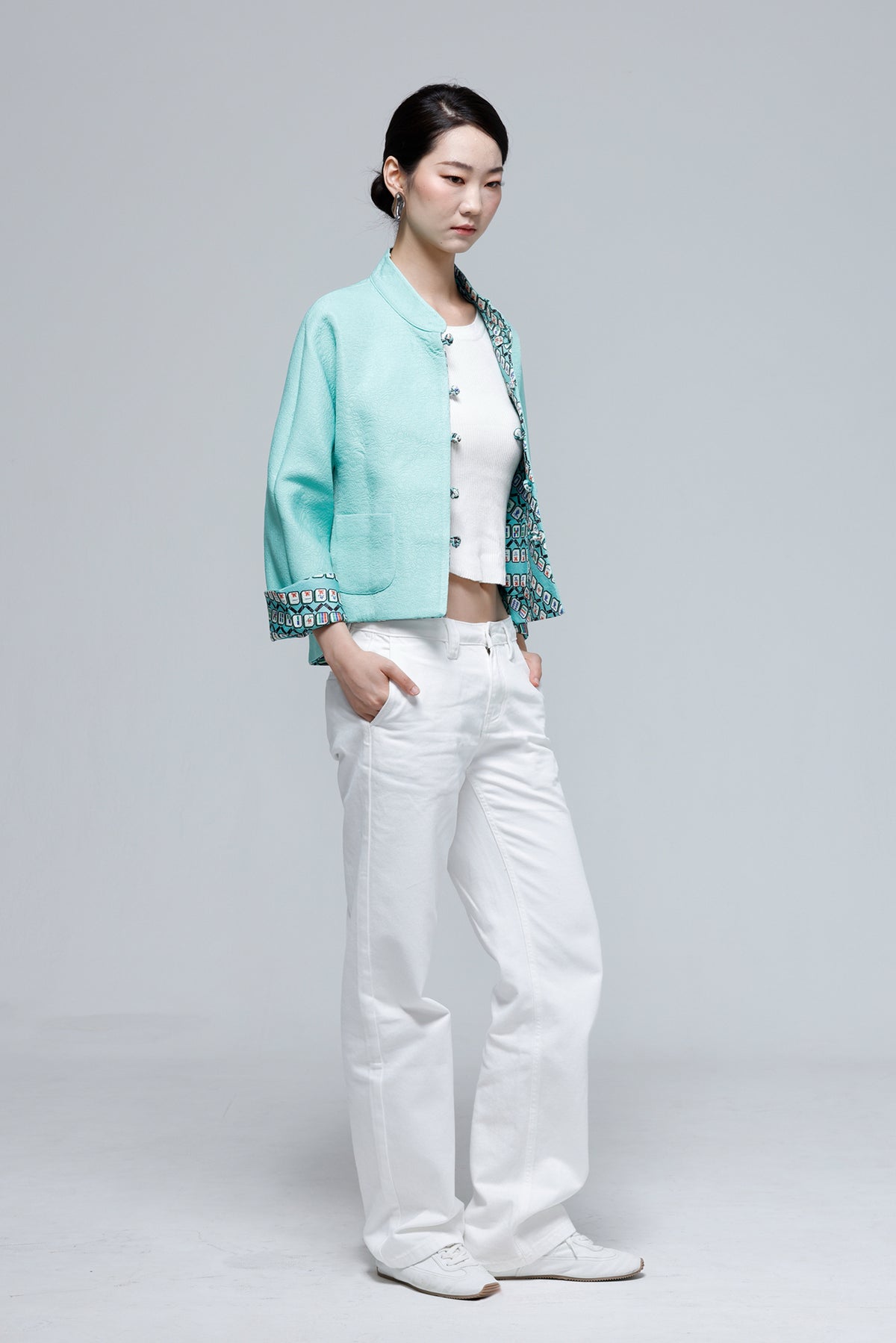 A woman modeling a light turquoise cropped jacket with patterned trim over a white top and white wide-leg jeans, standing against a light gray studio background.
