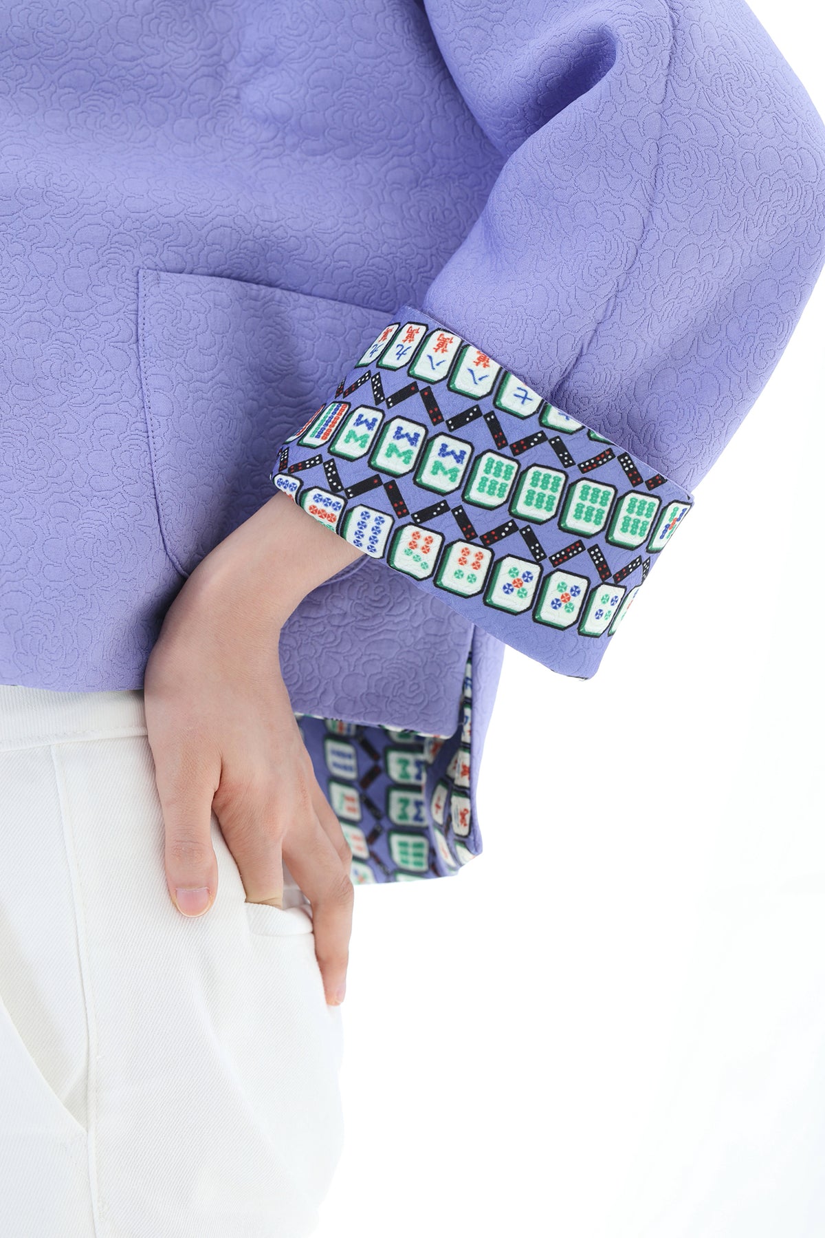 Close-up of a person wearing a lavender-purple textured jacket with a decorative multicolor patterned cuff, paired with white pants against a white background.