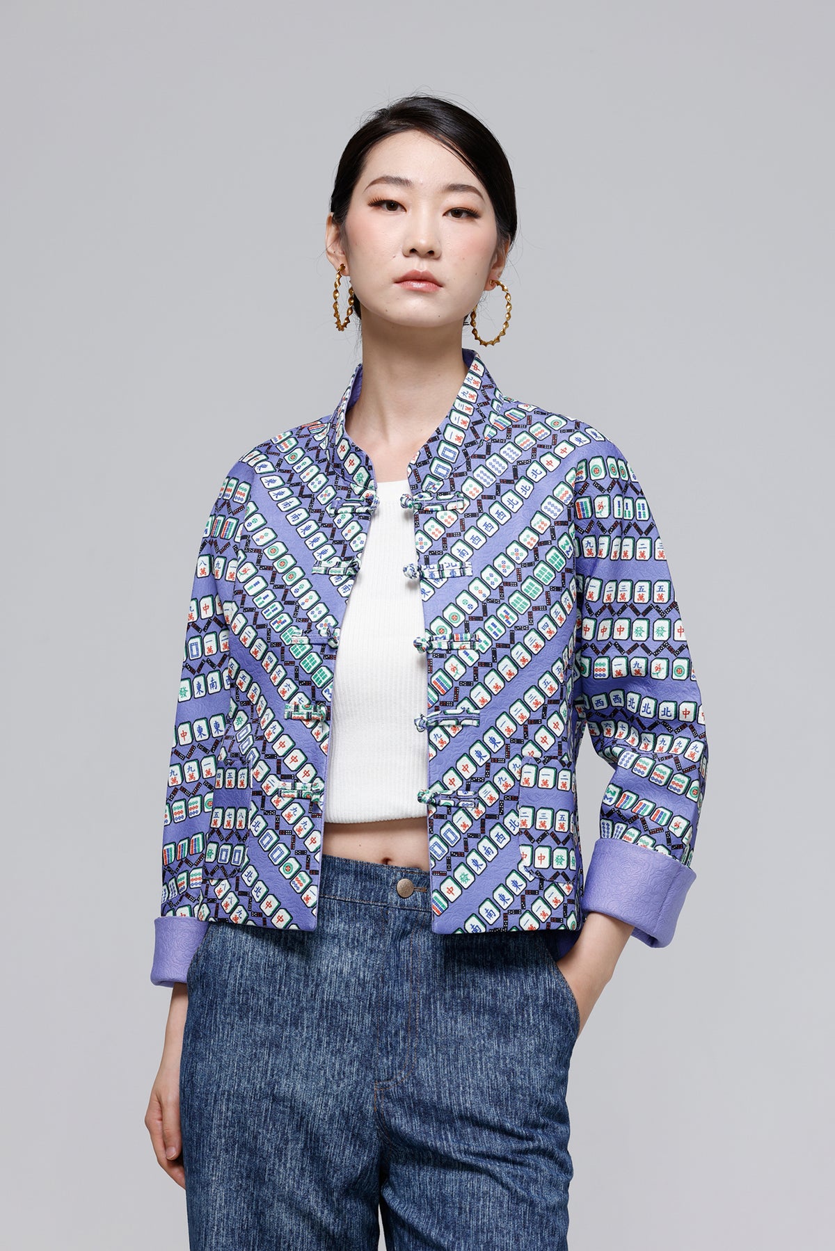 A woman modeling a short-sleeve cropped jacket with a geometric multicolor pattern in shades of purple, blue, green, and white, worn over a white top and blue jeans against a solid gray studio background.
