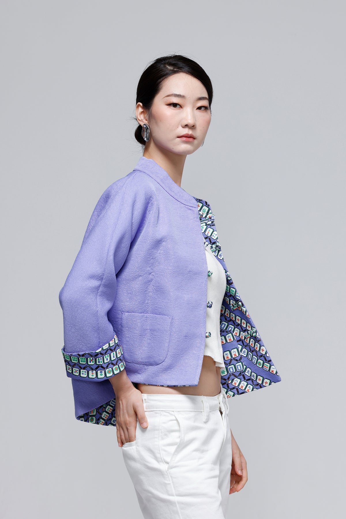 A model wearing a lavender cropped jacket with patterned trim, styled with white pants against a solid gray studio background.
