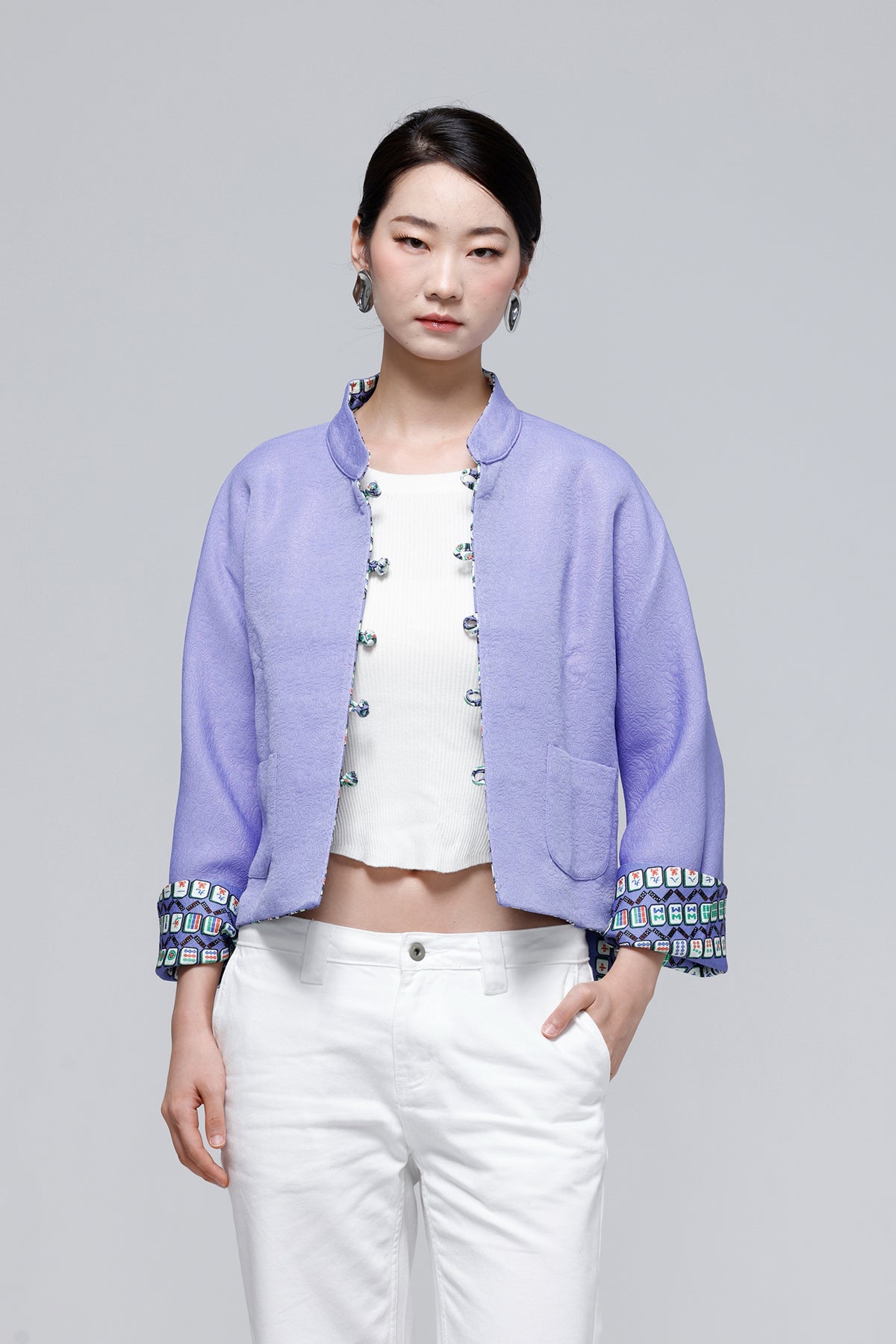 A woman modeling a light purple cropped jacket with colorful patterned trim on the cuffs and front, worn over a white crop top and white pants against a solid gray studio background.