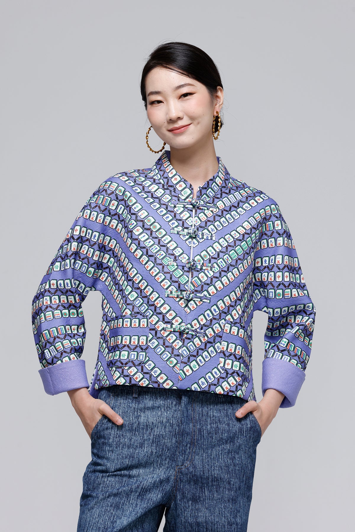 A woman modeling a long-sleeve button-up blouse with a geometric pattern in shades of purple, blue, and white, paired with blue jeans against a solid gray studio background.