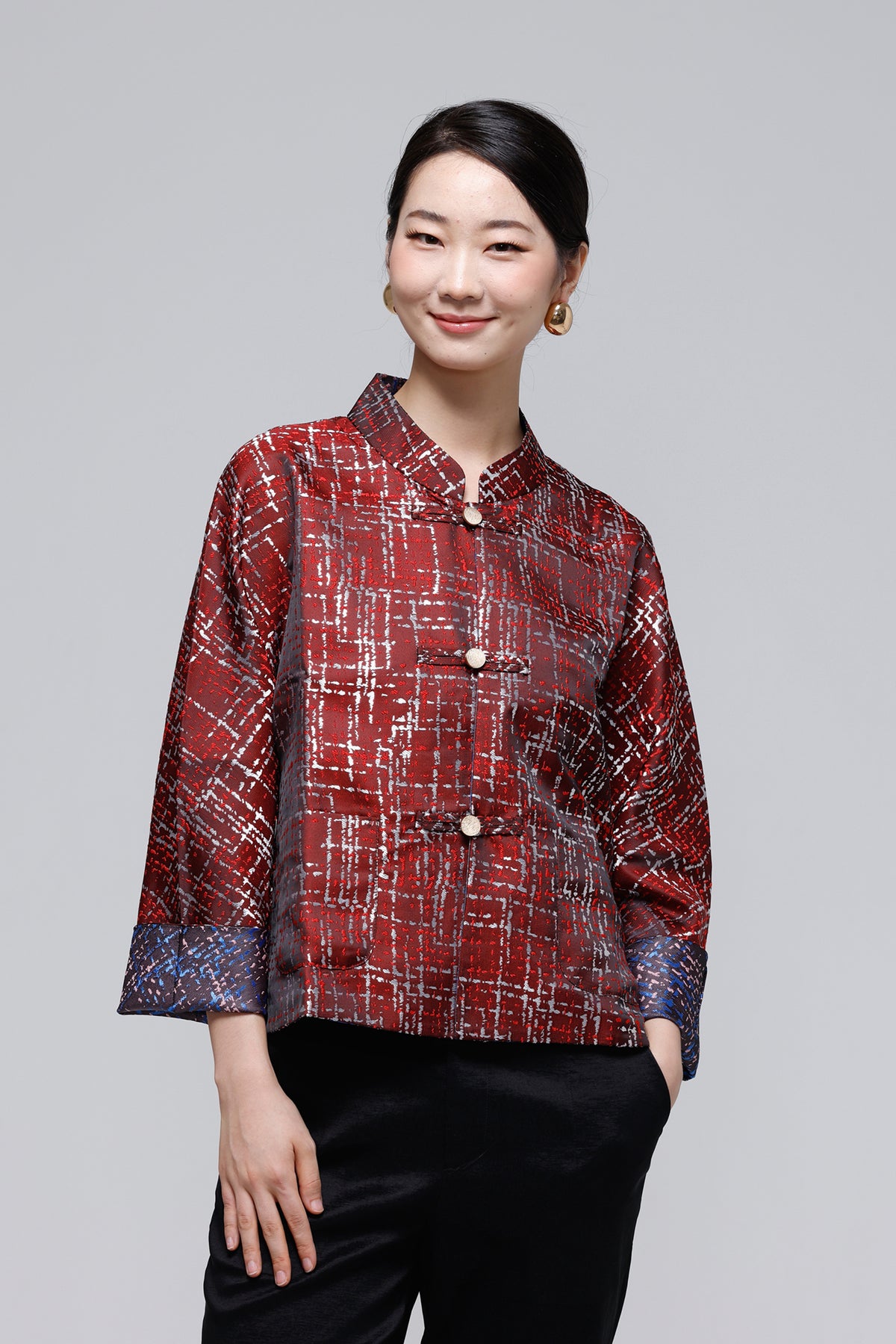 A woman modeling a red and black textured, plaid-style jacket with a mandarin collar and button closure, paired with black pants, against a solid gray studio background.