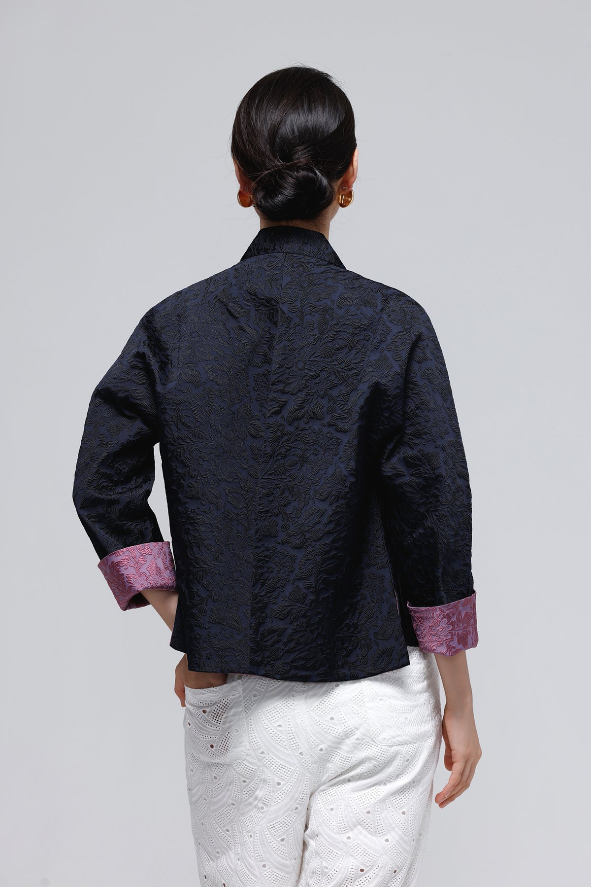 A woman shown from the back wearing a dark navy textured jacket with contrasting purple cuffs and white pants, set against a plain light gray studio background.