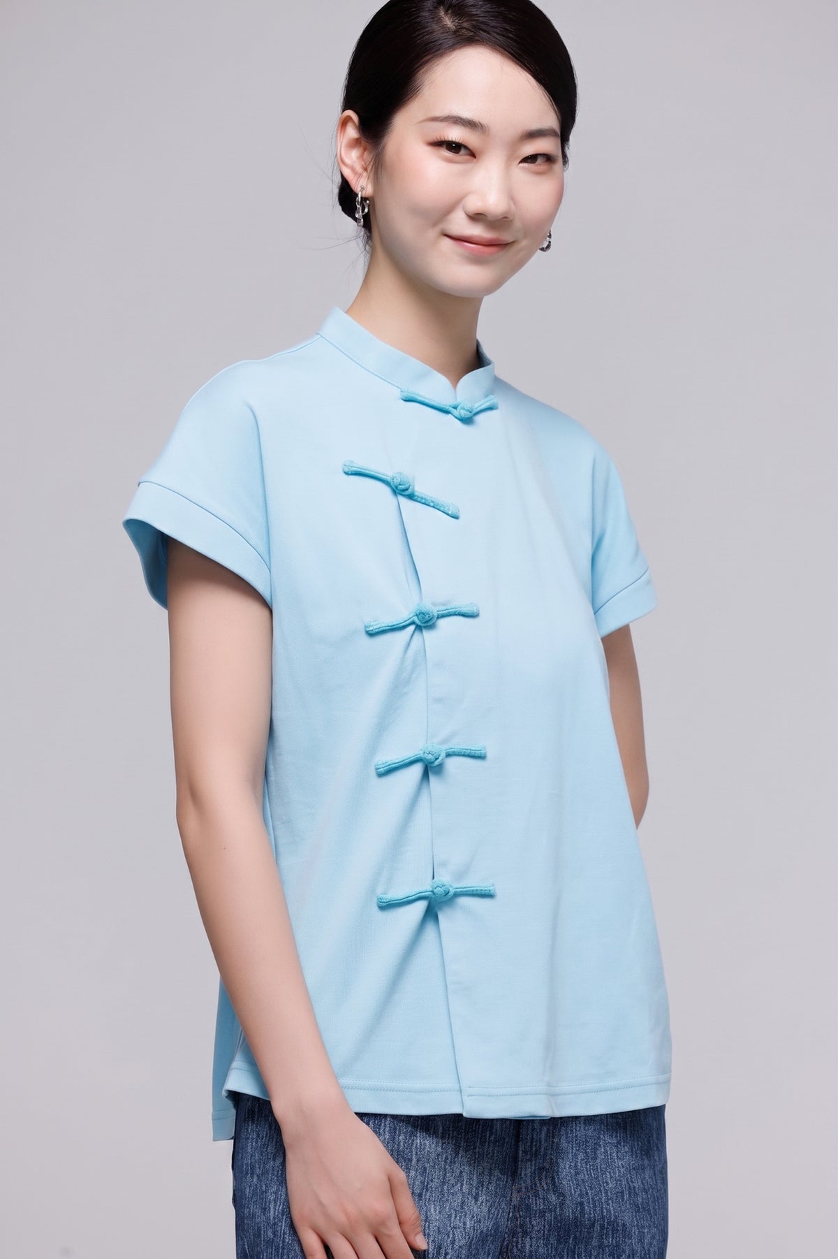 A woman modeling a light blue short-sleeve top with traditional Chinese-style frog closures, paired with blue jeans, against a solid gray studio background.
