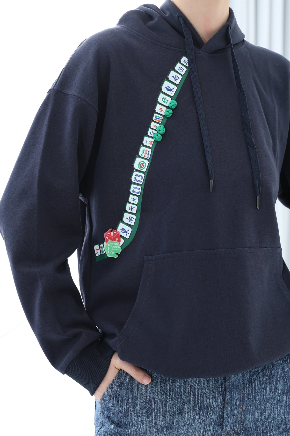 A person wearing a dark navy pullover hoodie with a vertical graphic print of green mahjong tiles and a red skull motif on the front, paired with blue jeans.