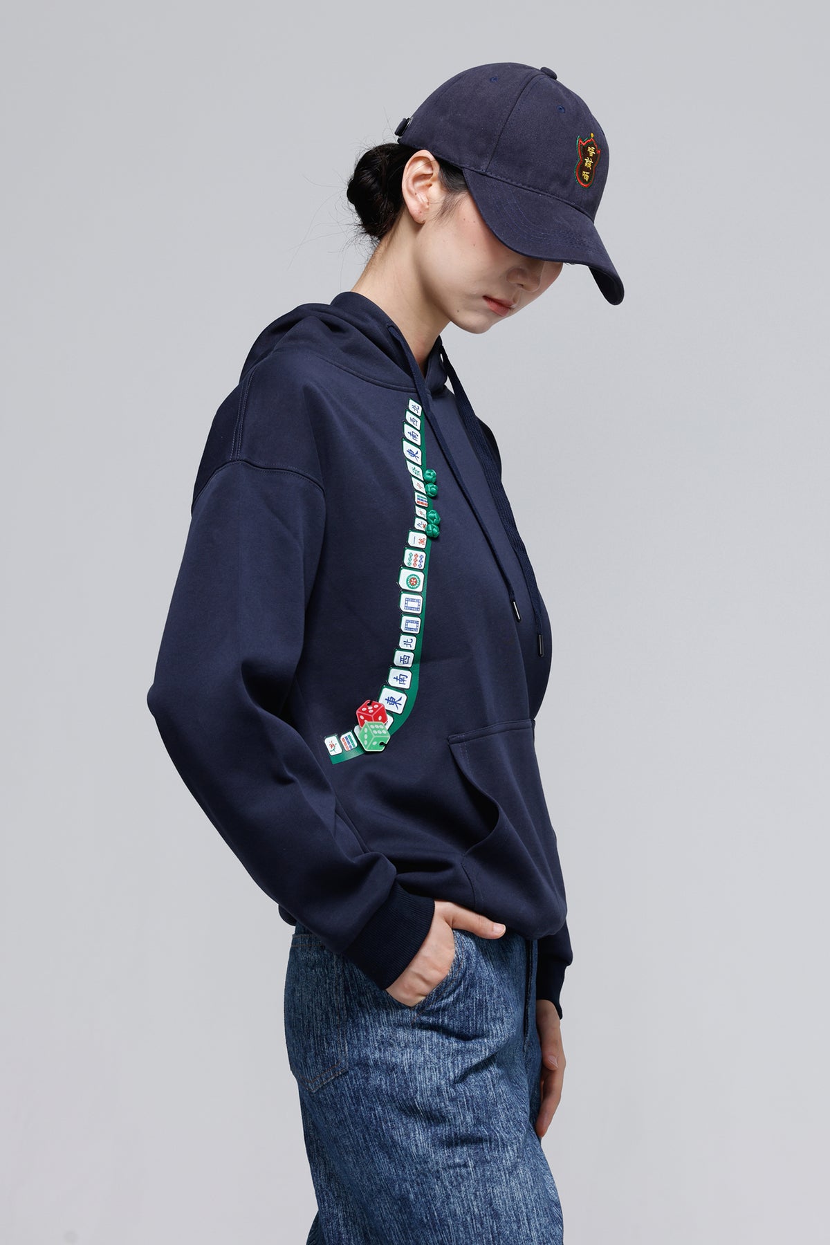 A model wearing a navy blue hoodie with a green and white checkered graphic down the left side, paired with a matching navy baseball cap and blue jeans, photographed against a neutral gray background.