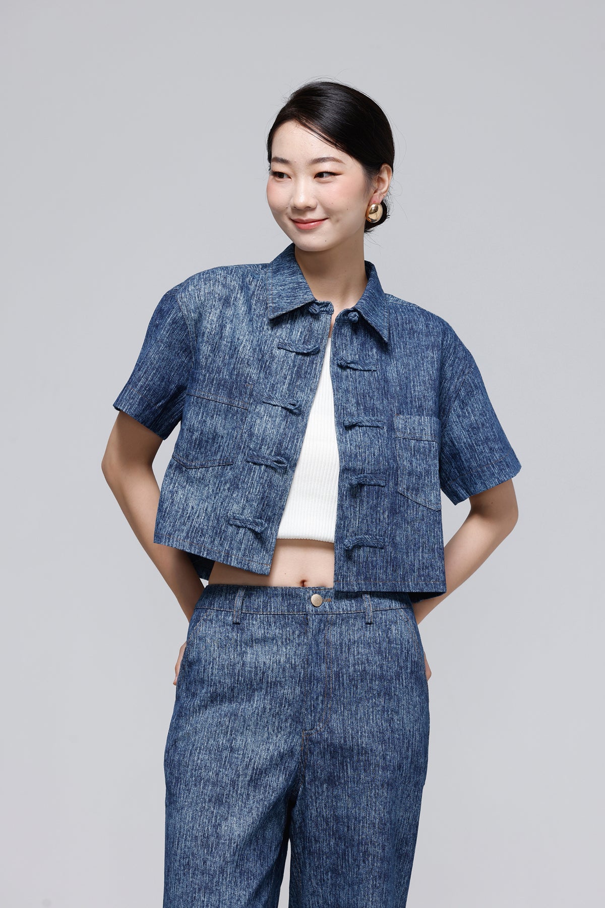 A woman modeling a blue textured short-sleeve cropped jacket with traditional Chinese frog closures, paired with matching blue jeans and a white top, against a solid gray studio background.