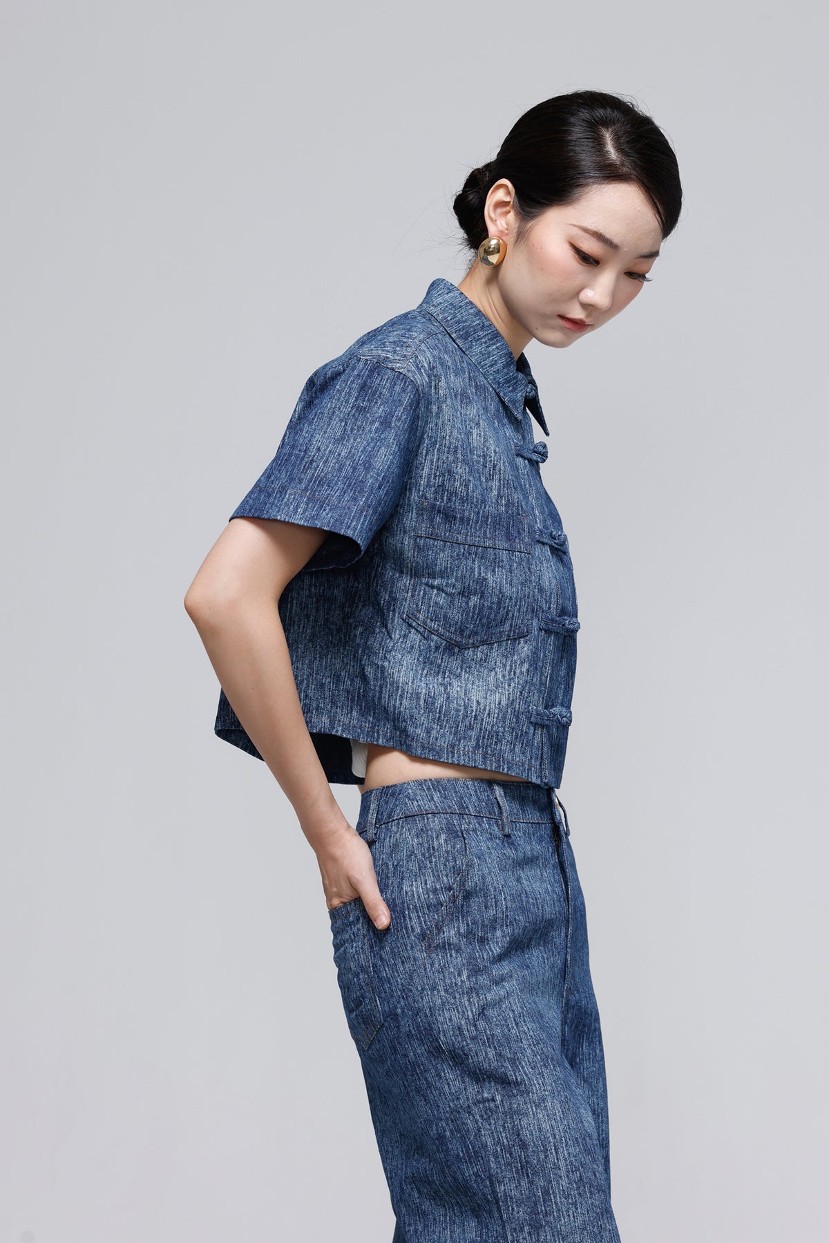 A woman modeling a matching blue denim short-sleeve cropped jacket and high-waisted wide-leg pants against a solid gray studio background.