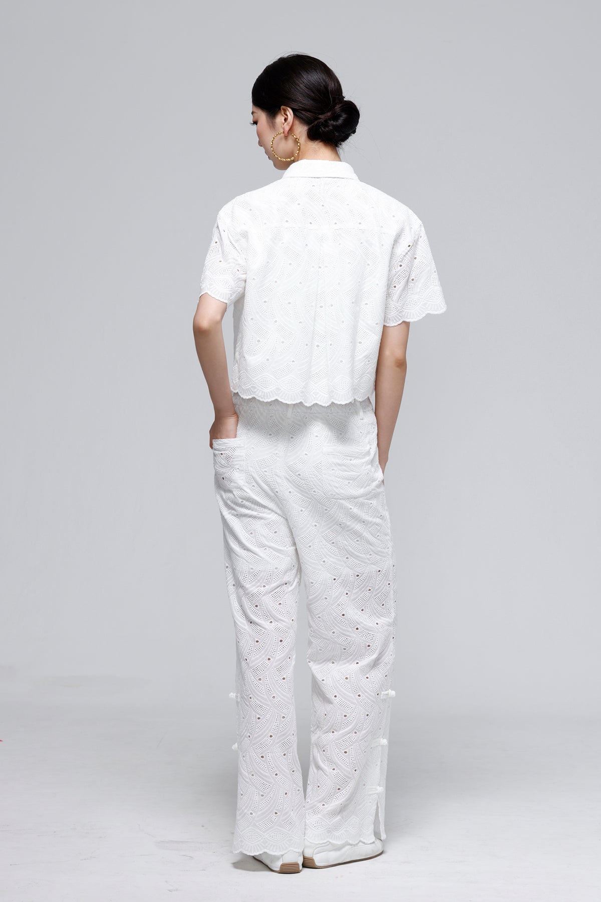 A woman shown from the back wearing a white short-sleeve cropped eyelet blouse and matching white wide-leg trousers against a light gray studio background.