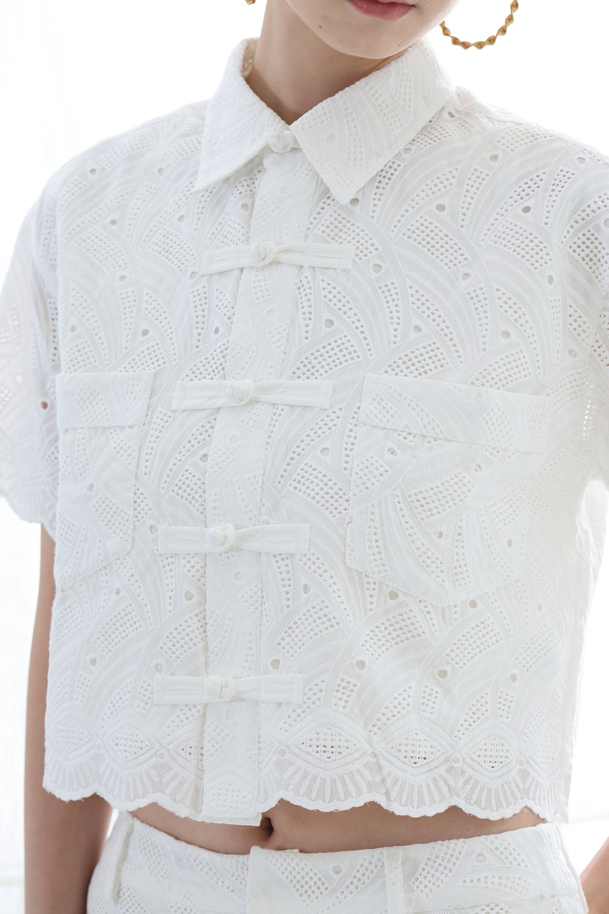 A close-up of a model wearing a white short-sleeve collared blouse with a scalloped eyelet lace pattern and front button closures, paired with matching white bottoms.