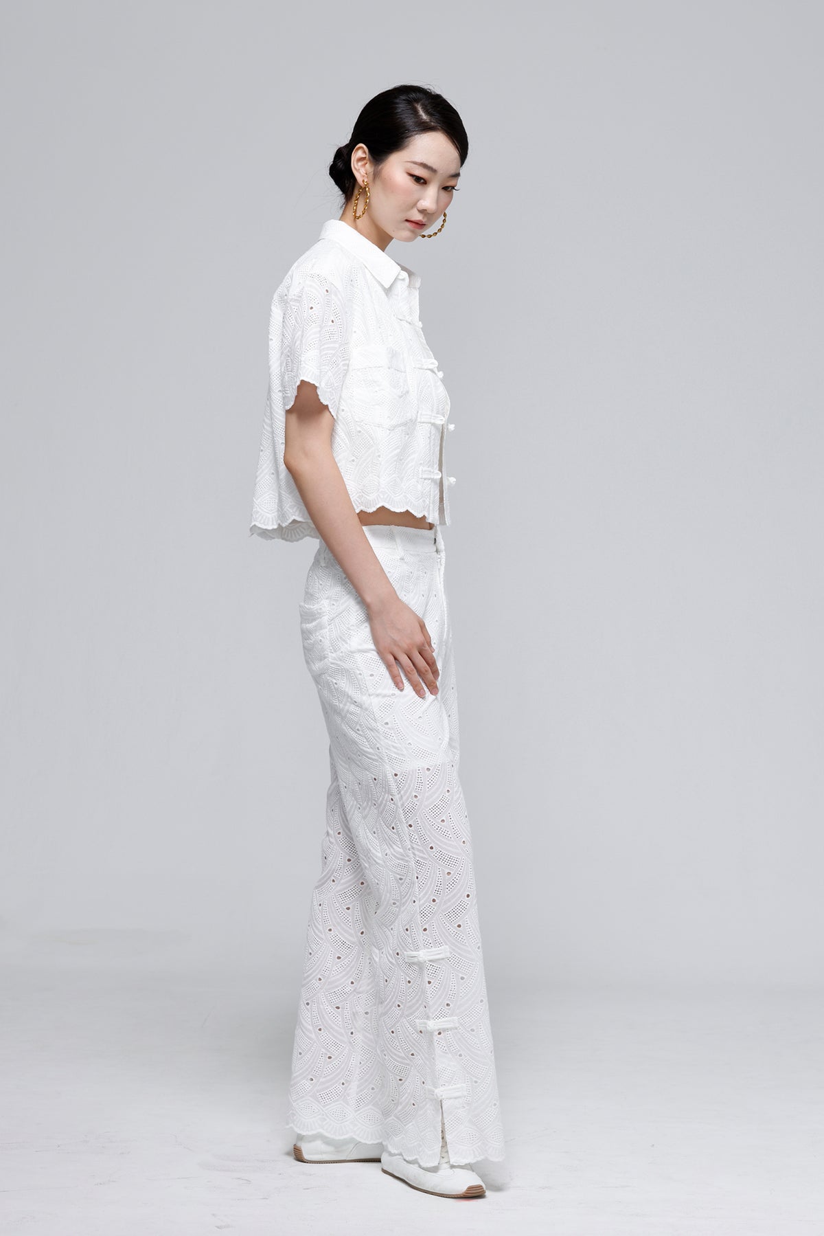 A woman modeling a white eyelet two-piece outfit consisting of a short-sleeve cropped button-up shirt and matching wide-leg pants, photographed against a light gray studio background.