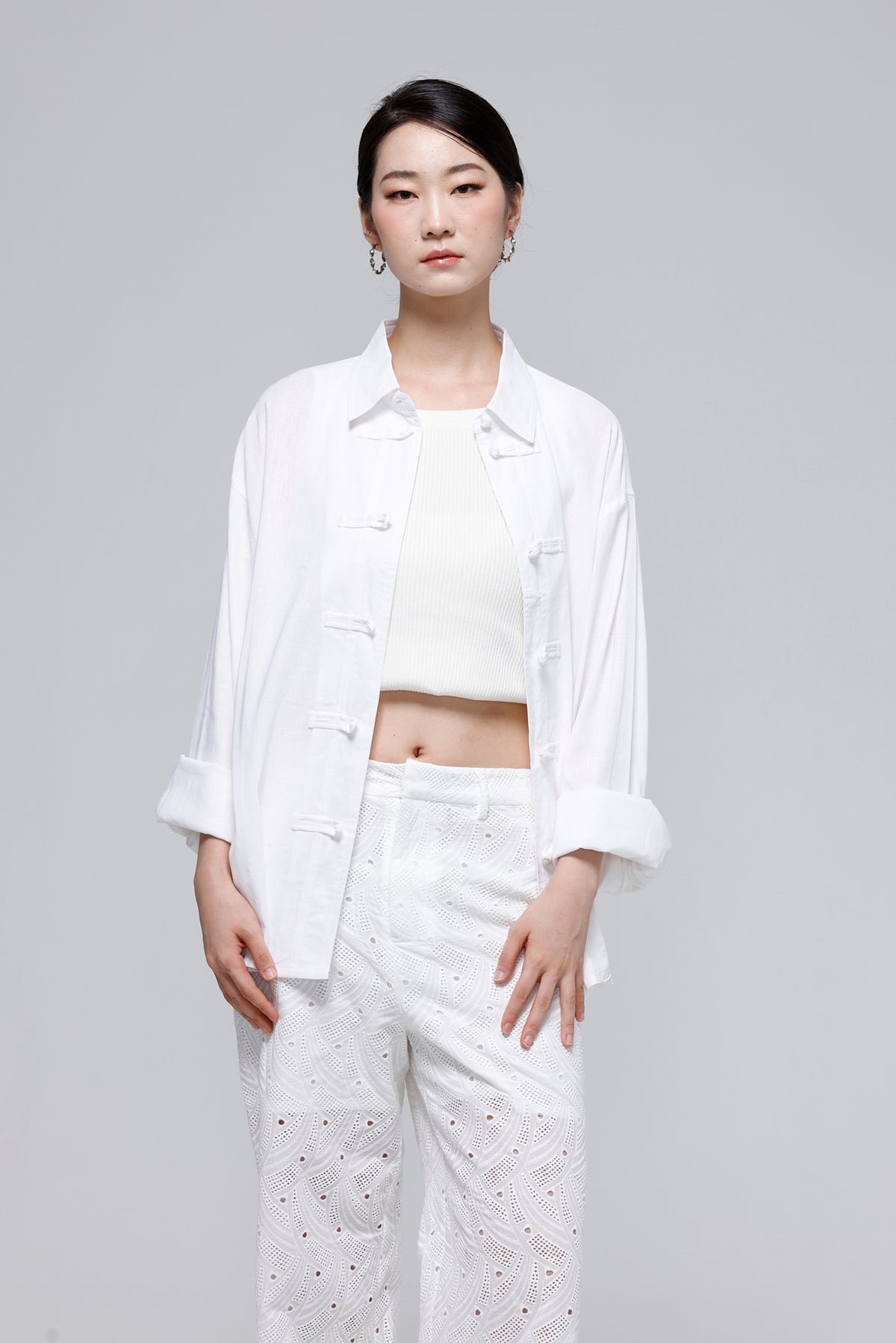 A model wearing a white three-piece outfit consisting of a long-sleeve button-front overshirt, a cropped tank top, and white eyelet trousers, shot against a solid light gray background.
