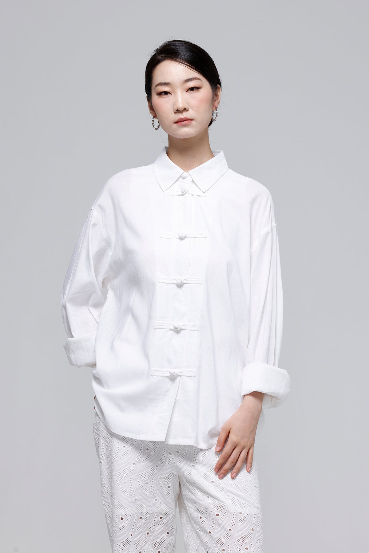 A woman modeling a white long-sleeve button-front blouse with a pointed collar and white textured pants against a light gray studio background.