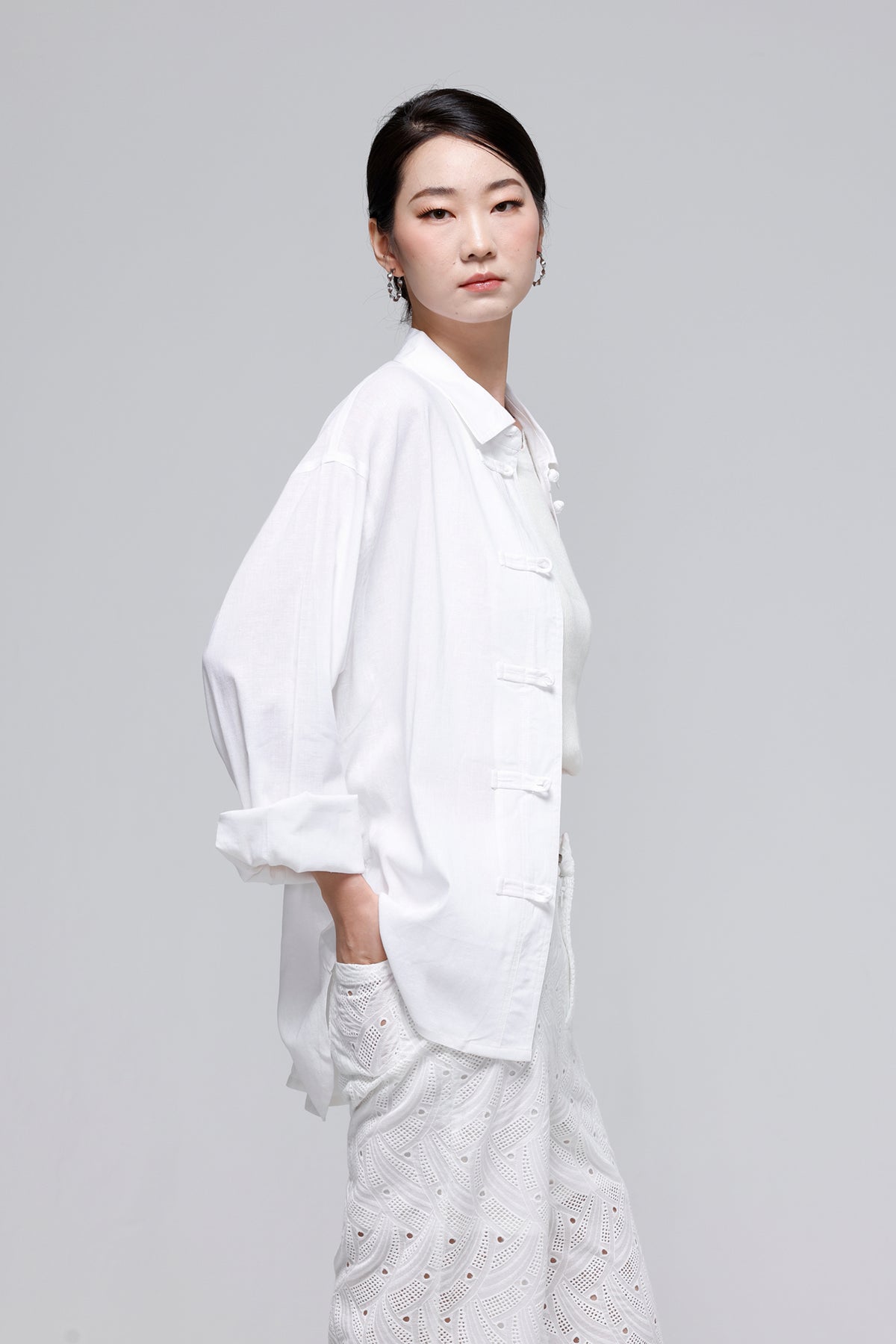 A woman modeling a white long-sleeve button-front shirt with rolled cuffs and a matching white eyelet skirt against a light gray studio background.