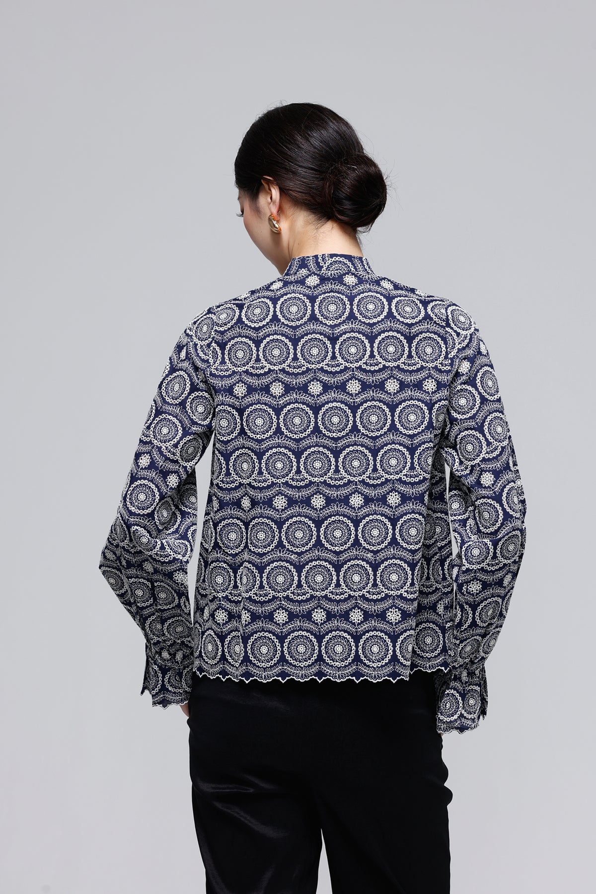 A woman viewed from the back wearing a long-sleeve blouse with a navy blue and white circular mandala pattern, paired with black pants, against a solid light gray background.