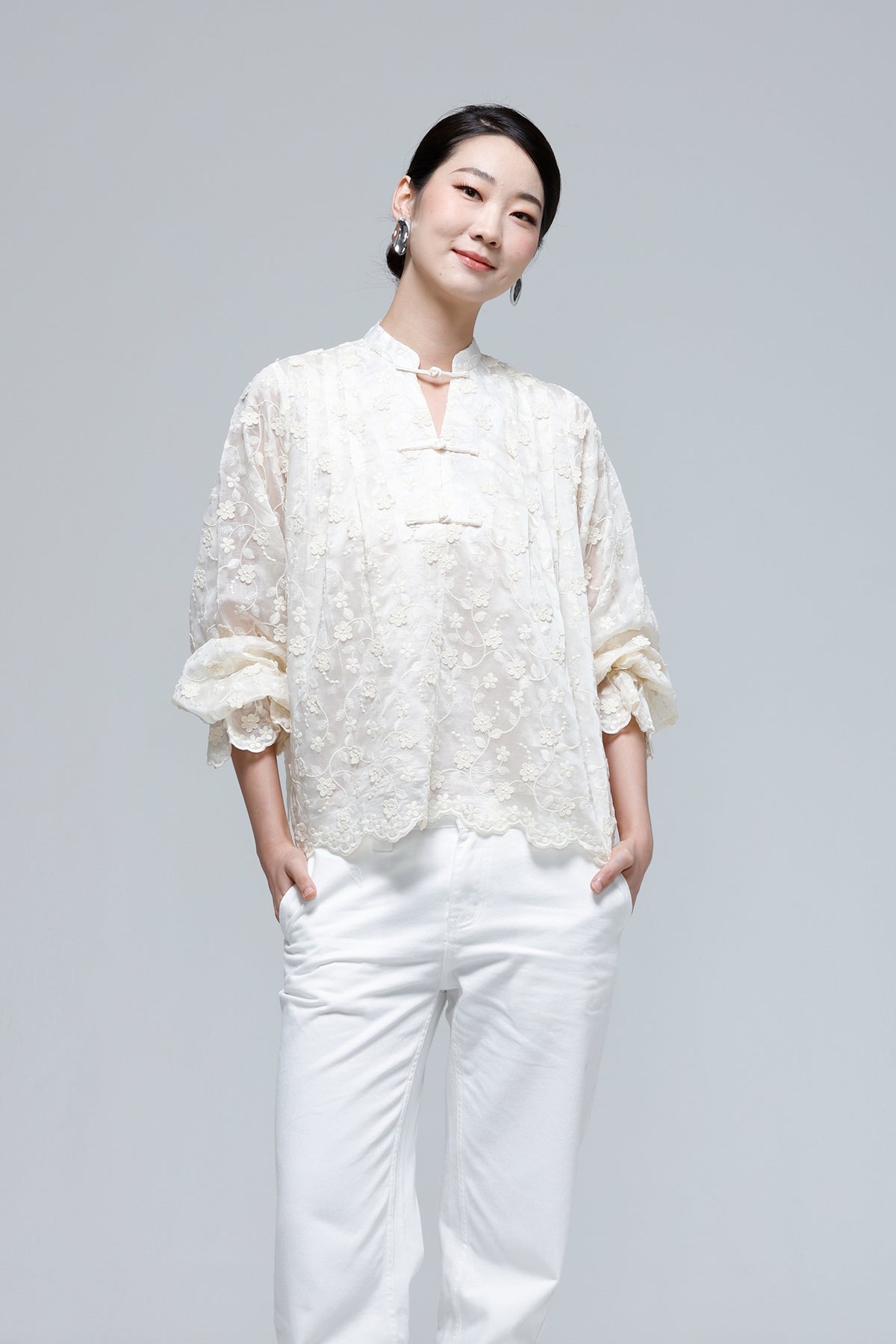 A woman modeling a cream-colored, semi-sheer floral lace blouse with traditional Chinese-style collar and frog closures, paired with white trousers, against a solid light gray background.