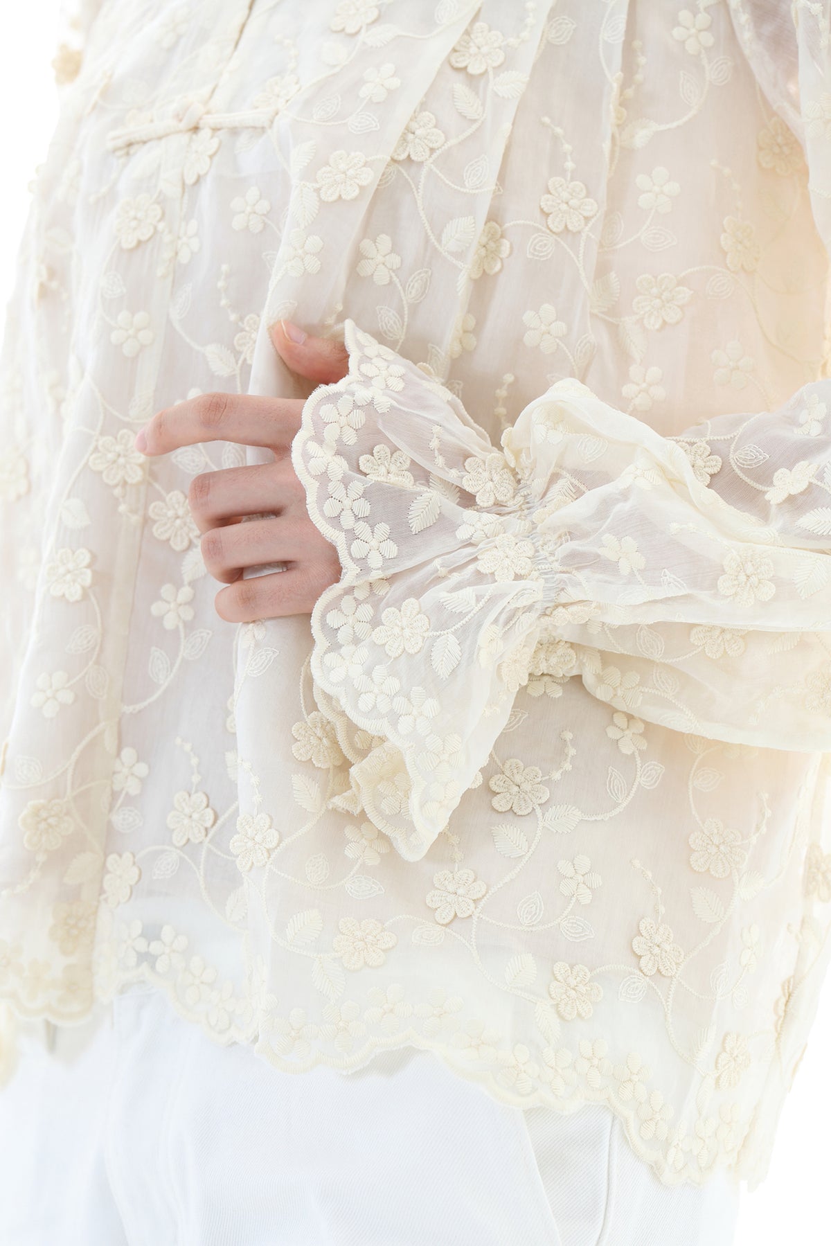 Close-up of a cream-colored sheer embroidered blouse with floral lace detailing and a ruffled long sleeve, worn by a model.
