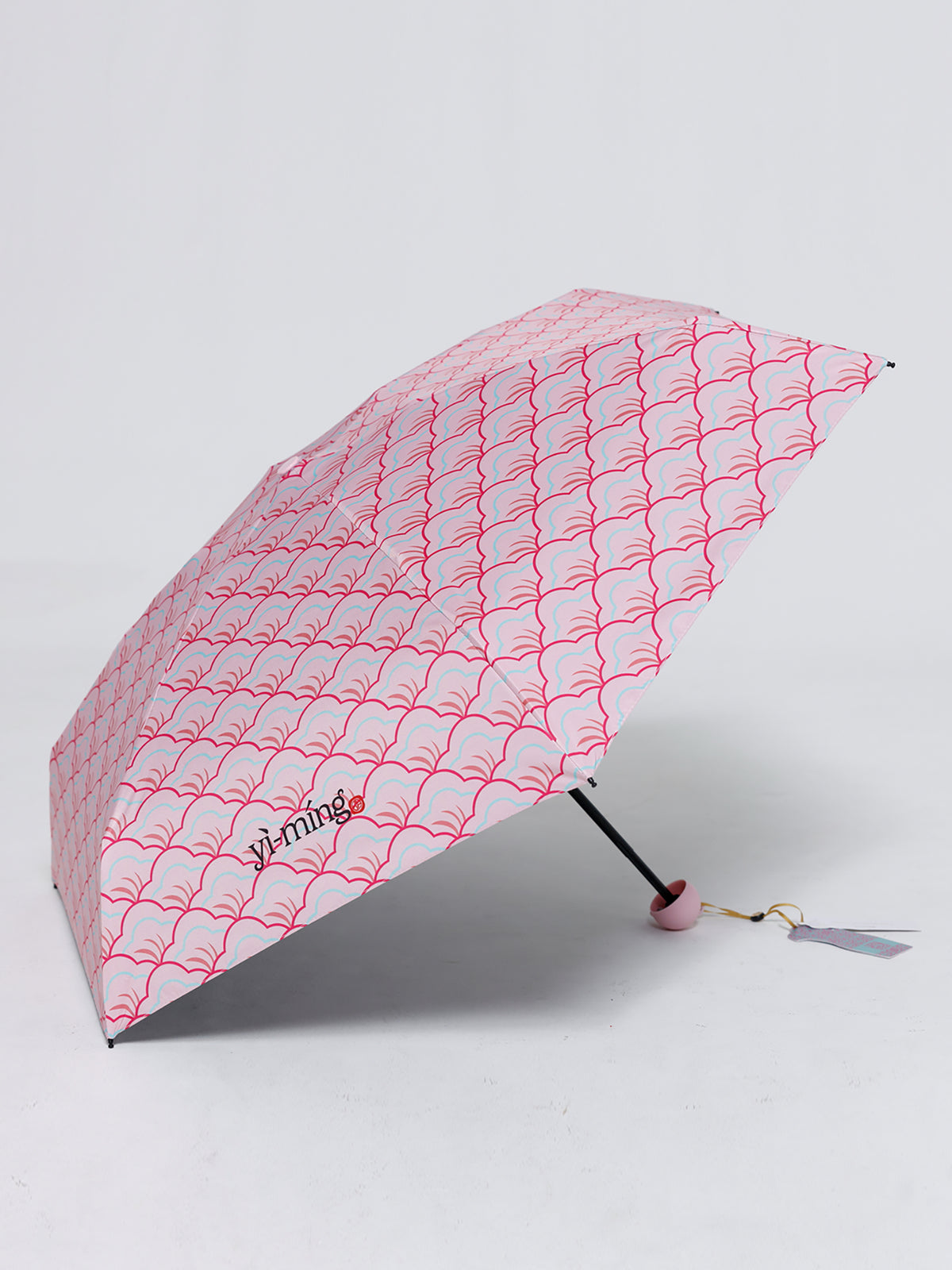 A compact folding umbrella with a pink  repeating wave pattern, shown open on a light gray studio background. The brand name 'yi-ming' is printed in black near the lower left of the canopy.
