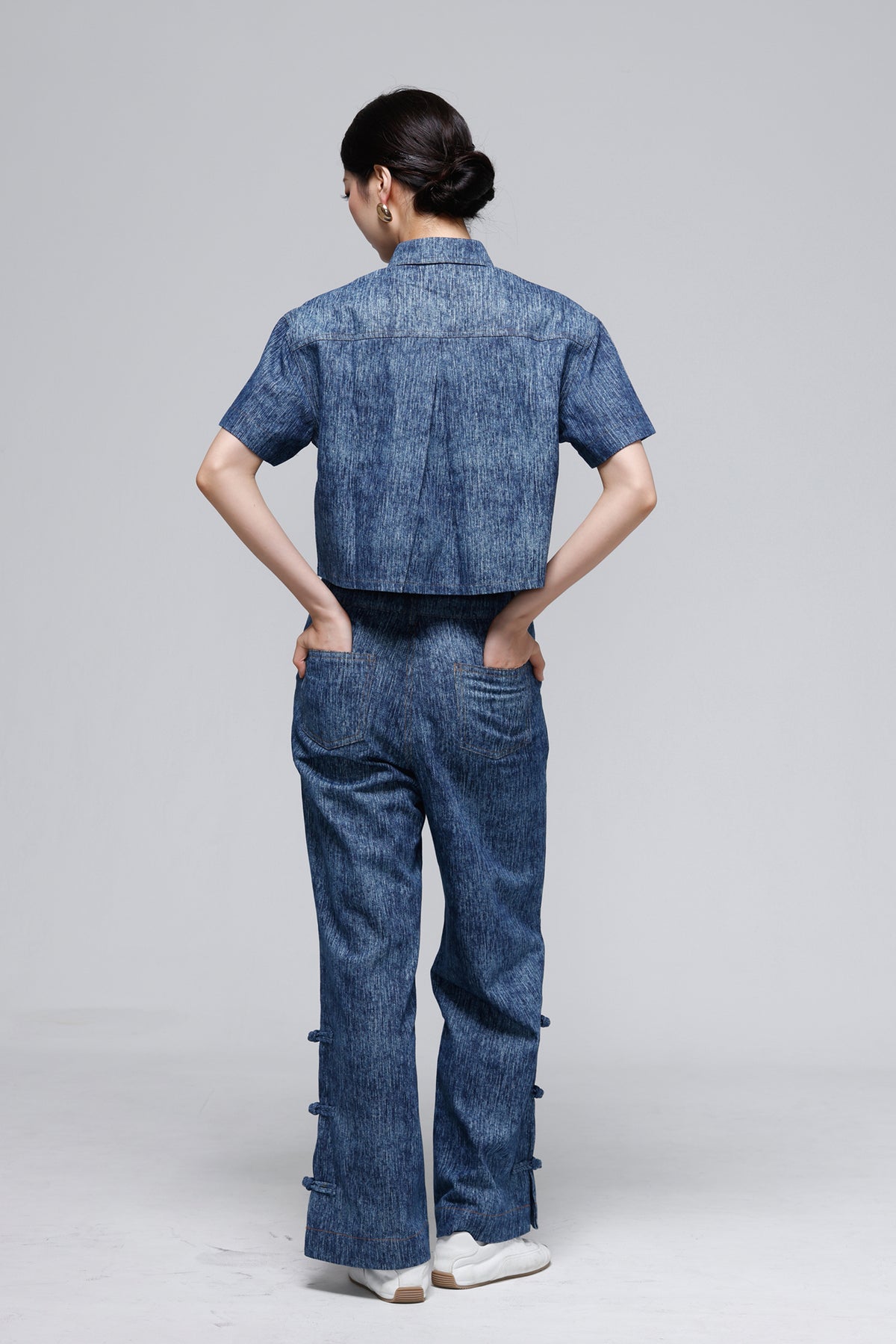 A model viewed from the back wearing a matching blue denim short-sleeve top and wide-leg pants set, standing against a light gray studio background.