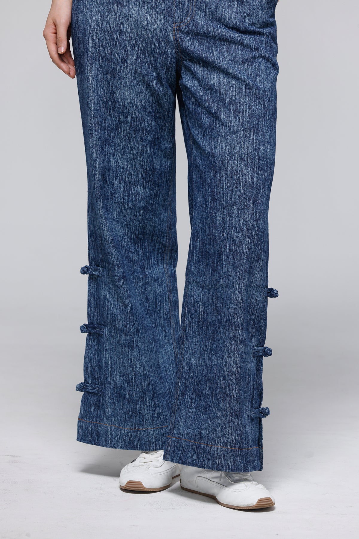 A person wearing blue wide-leg denim pants with decorative dark blue fabric ties along the outer sides, paired with white sneakers, photographed against a light gray studio background.