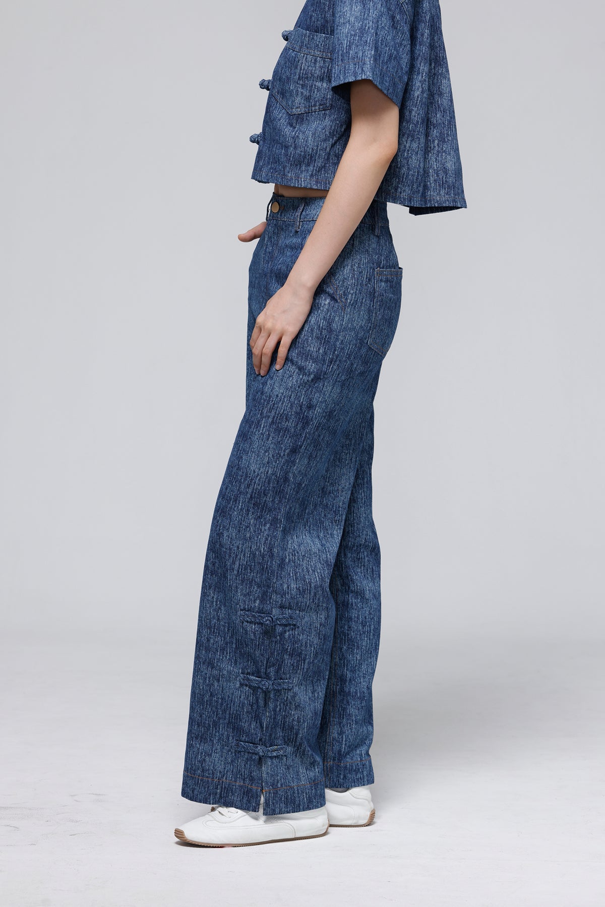 A model shown from the side wearing a matching blue denim set consisting of a cropped short-sleeve top with decorative buttons and high-waisted, wide-leg pants with front patch pockets, styled with white sneakers against a light gray studio background.