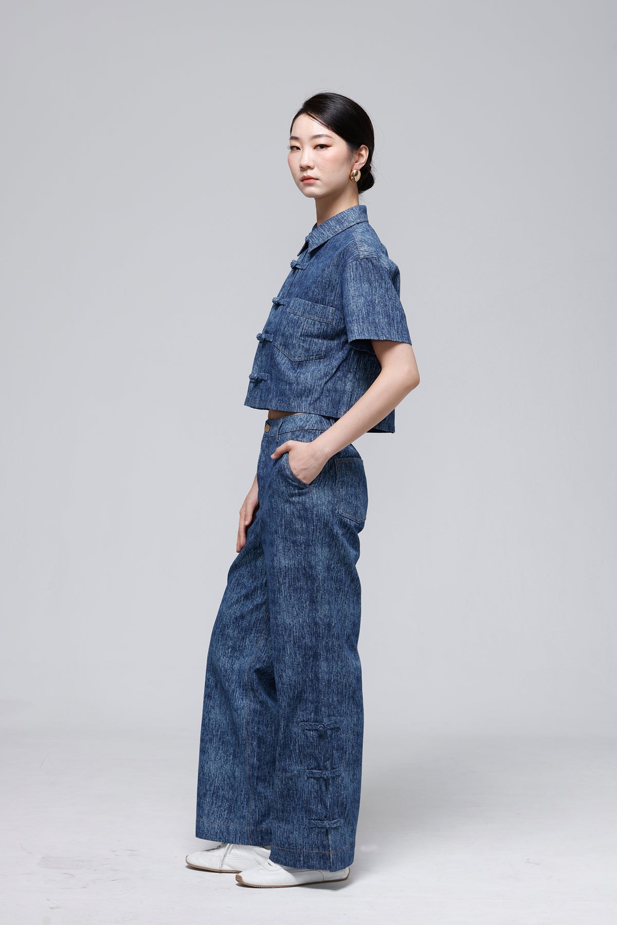 A model wearing a matching blue denim two-piece outfit consisting of a short-sleeve cropped top and wide-leg pants, standing in profile against a light gray studio background.