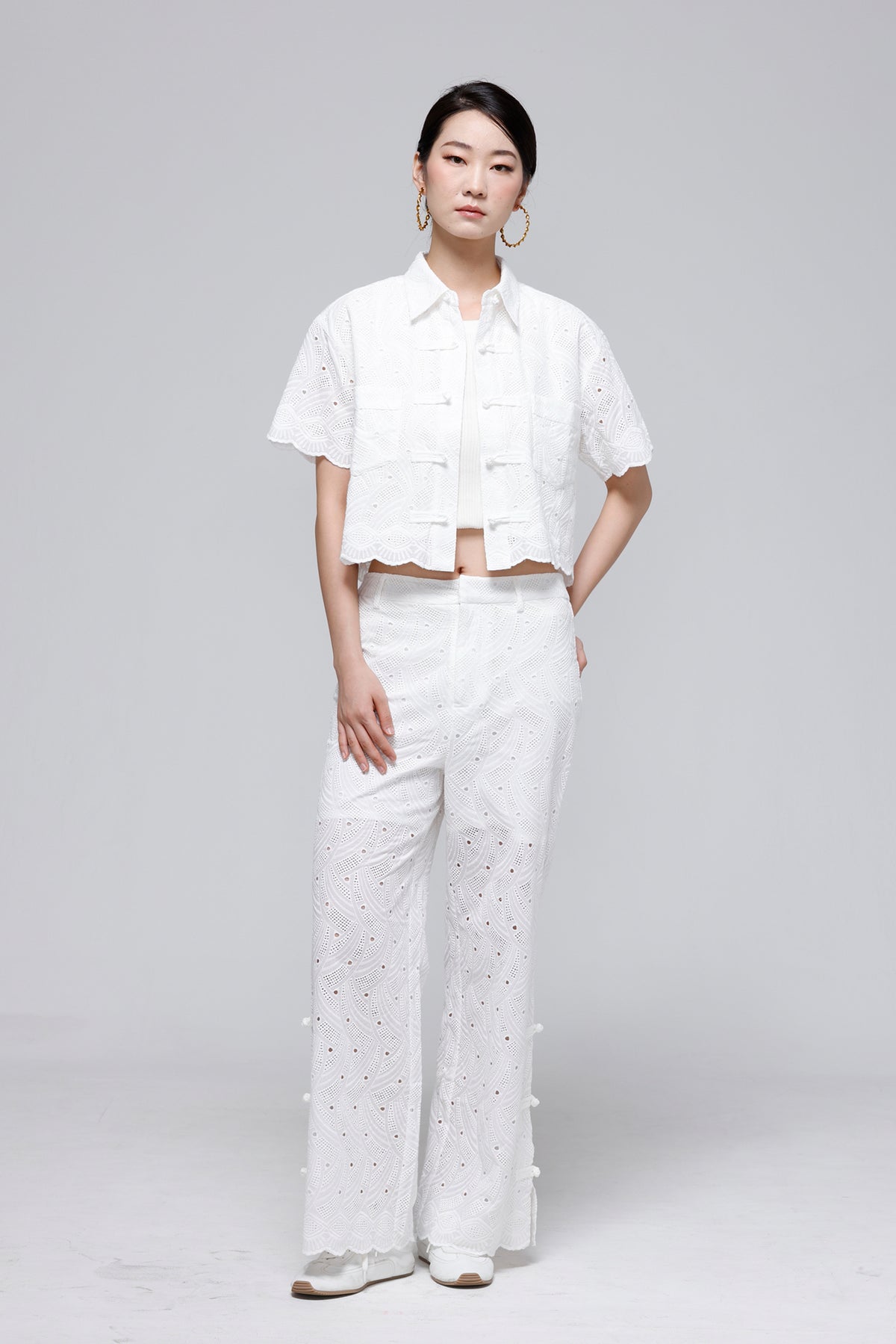 A model stands against a light gray studio background wearing a white short-sleeve cropped eyelet blouse and matching white wide-leg eyelet trousers with decorative buttons down the side.