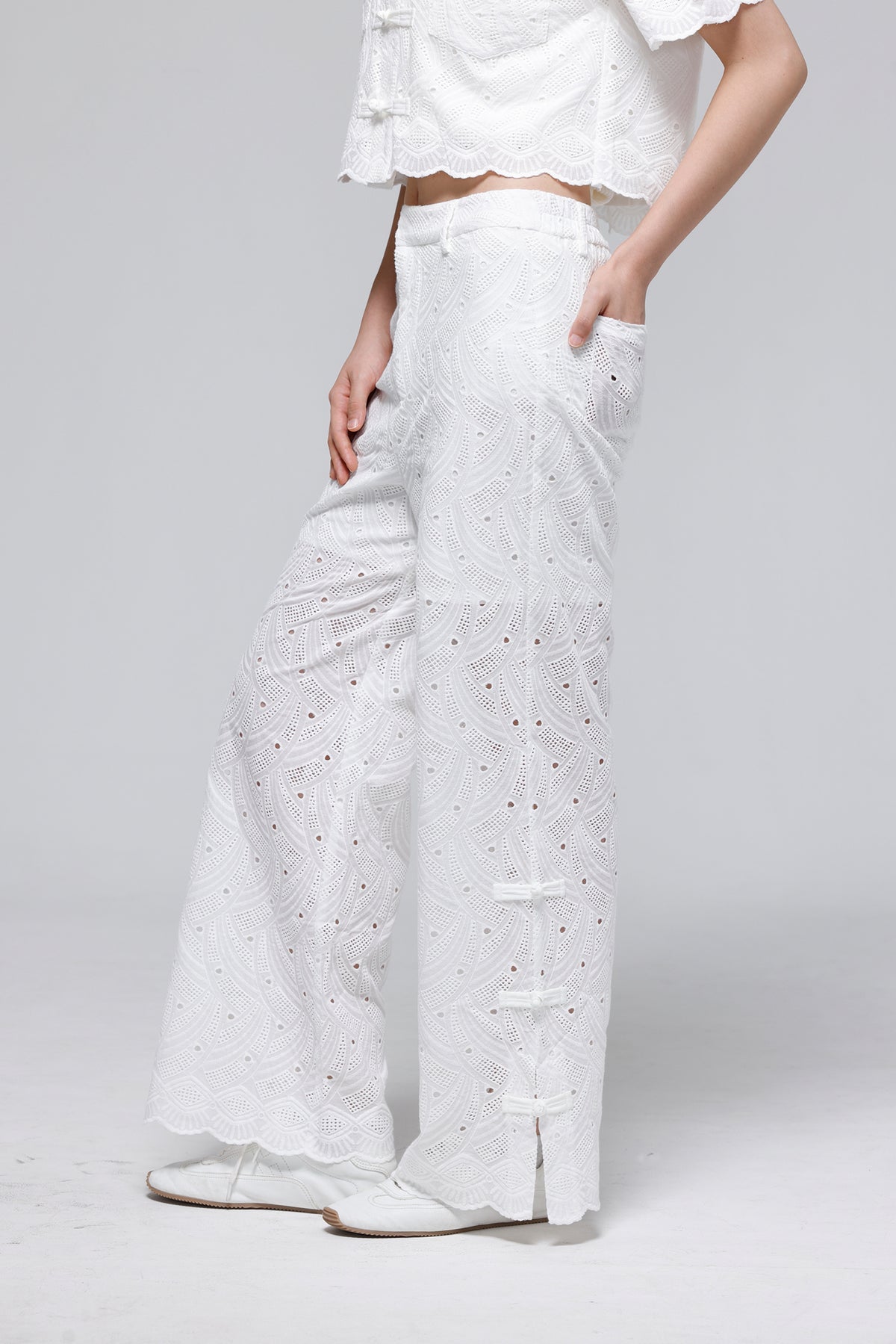 White eyelet lace wide-leg pants with side button closures and a scalloped hem, shown on a model against a light gray studio background.