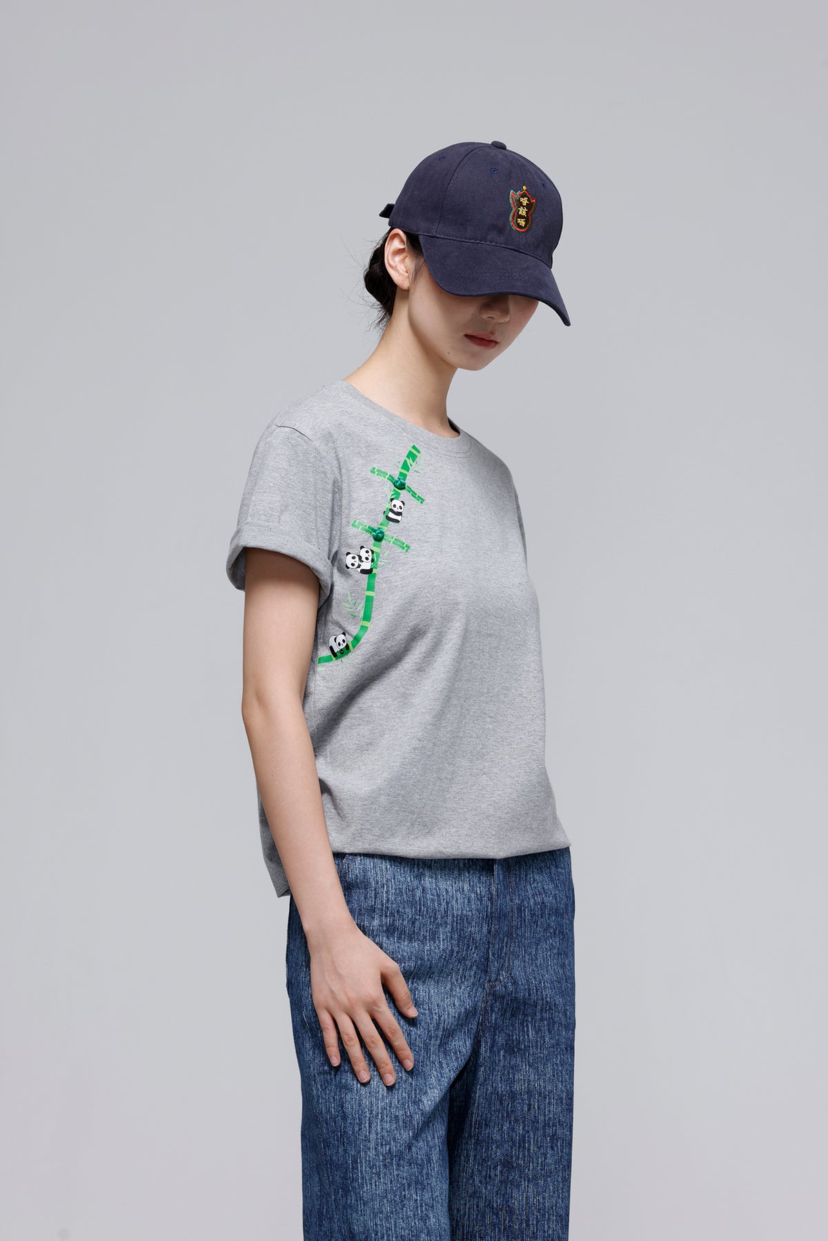 A model wearing a light gray crewneck t-shirt with a green bamboo graphic and panda print on the front, paired with blue jeans and a navy blue baseball cap, set against a plain gray studio background.
