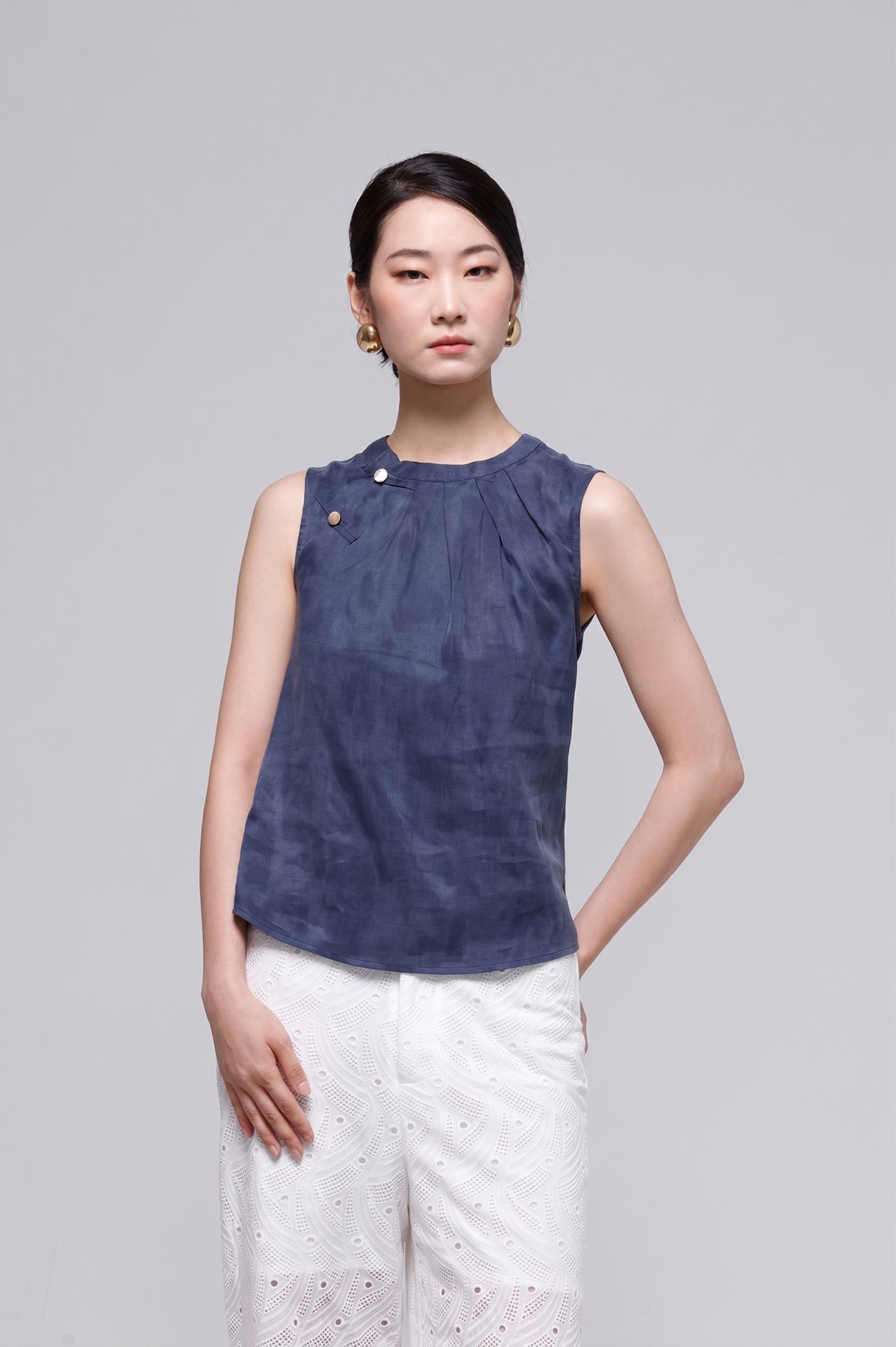 A woman modeling a sleeveless navy blue top with a gathered neckline and white pants, standing against a solid light gray studio background.