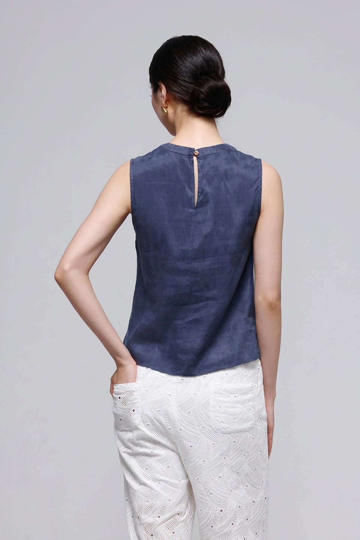 A woman shown from the back wearing a sleeveless navy blue top with a small back slit and white textured pants, photographed against a plain light gray studio background.